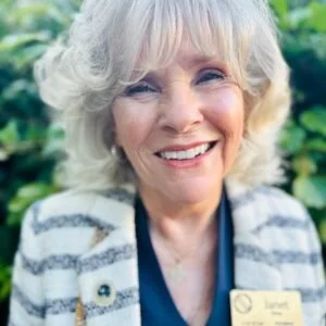 Smiling elderly woman with gray hair and blue eyes, wearing a striped cardigan and name tag, outdoors with greenery in the background.