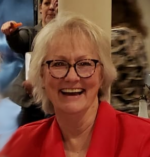 Smiling woman with glasses and blonde hair, wearing a red blazer, in an indoor setting with blurred background.