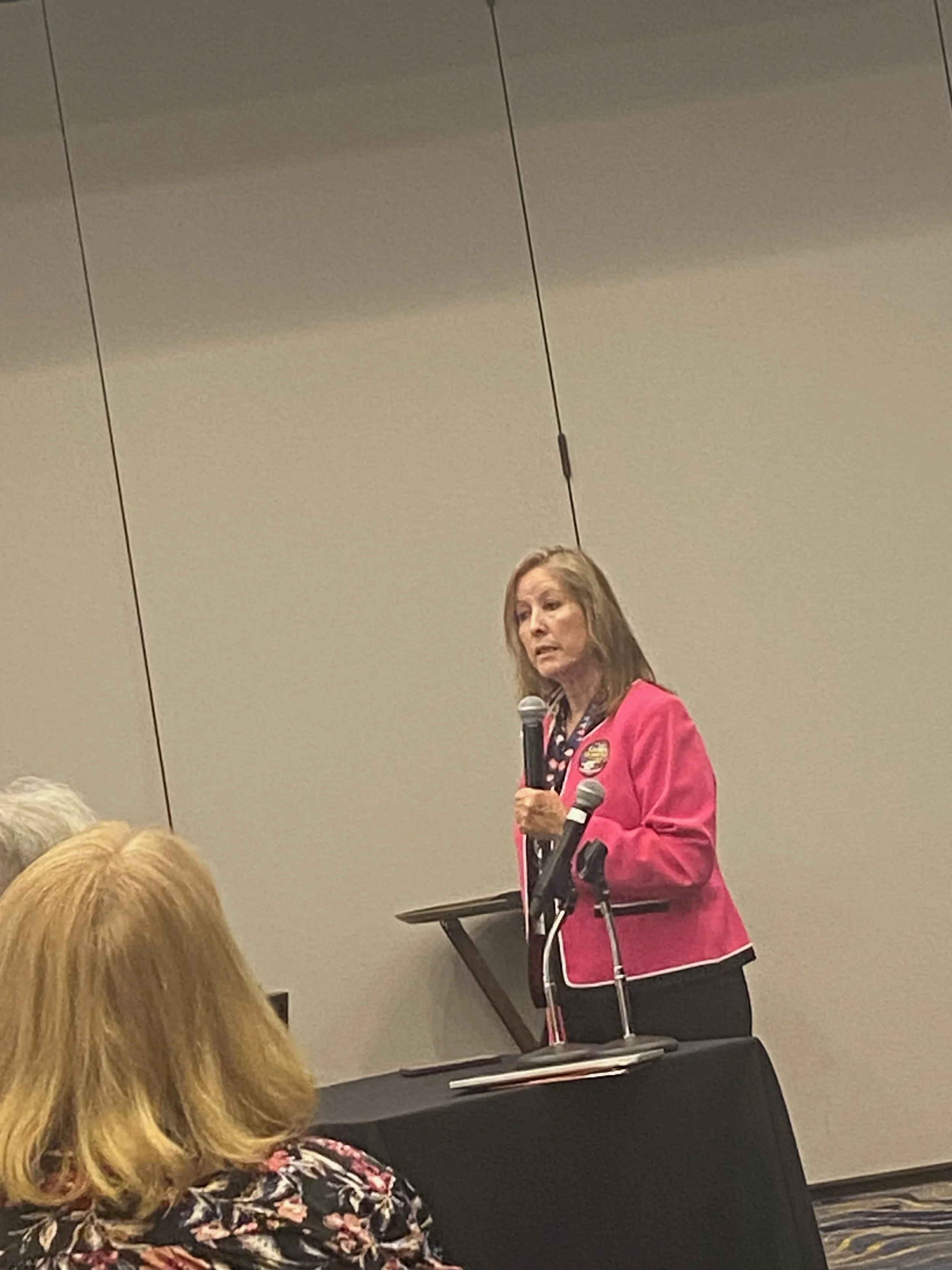 A woman with long hair, wearing a red jacket, standing at a podium, speaking into a microphone, in front of an audience in a conference room.