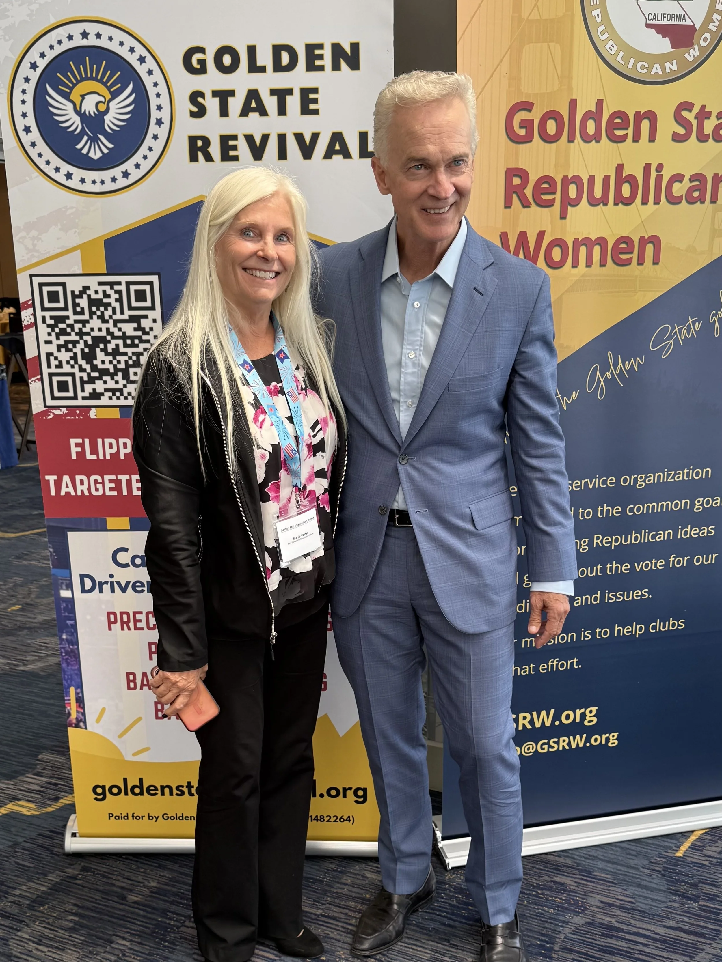 A woman and a man standing side by side at an event, smiling for the camera in front of promotional signs for Golden State Revival and Golden State Republican Women. The woman has long blond hair and is dressed in black pants and a colorful blouse, a