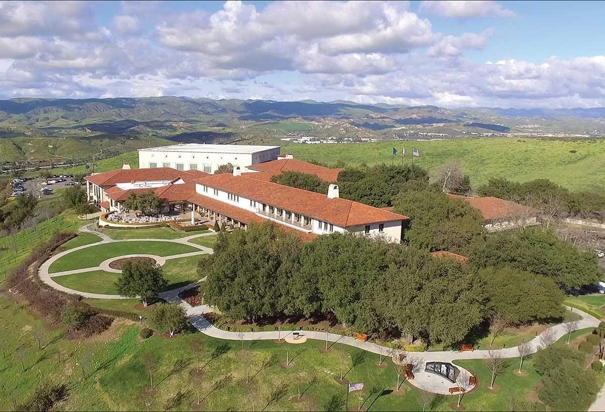 Aerial view of a large building with a red-tiled roof, surrounded by well-maintained green lawns, trees, and winding pathways. In the background, rolling hills and a cloudy sky are visible.