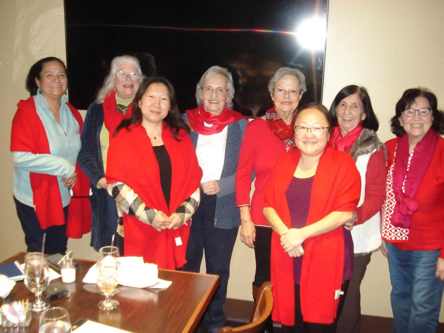 Group of ten women gathered around a table at a celebration or event, all wearing red or red accessories, smiling and posing for the photo indoors.