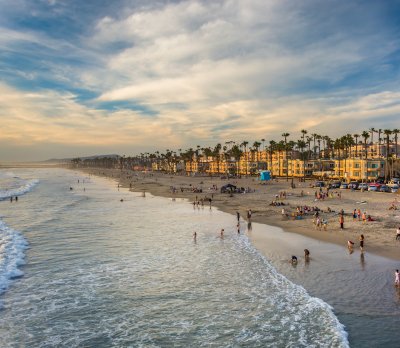 Beach with people swimming and relaxing, with hotels and palm trees in the background, under a partly cloudy sky.