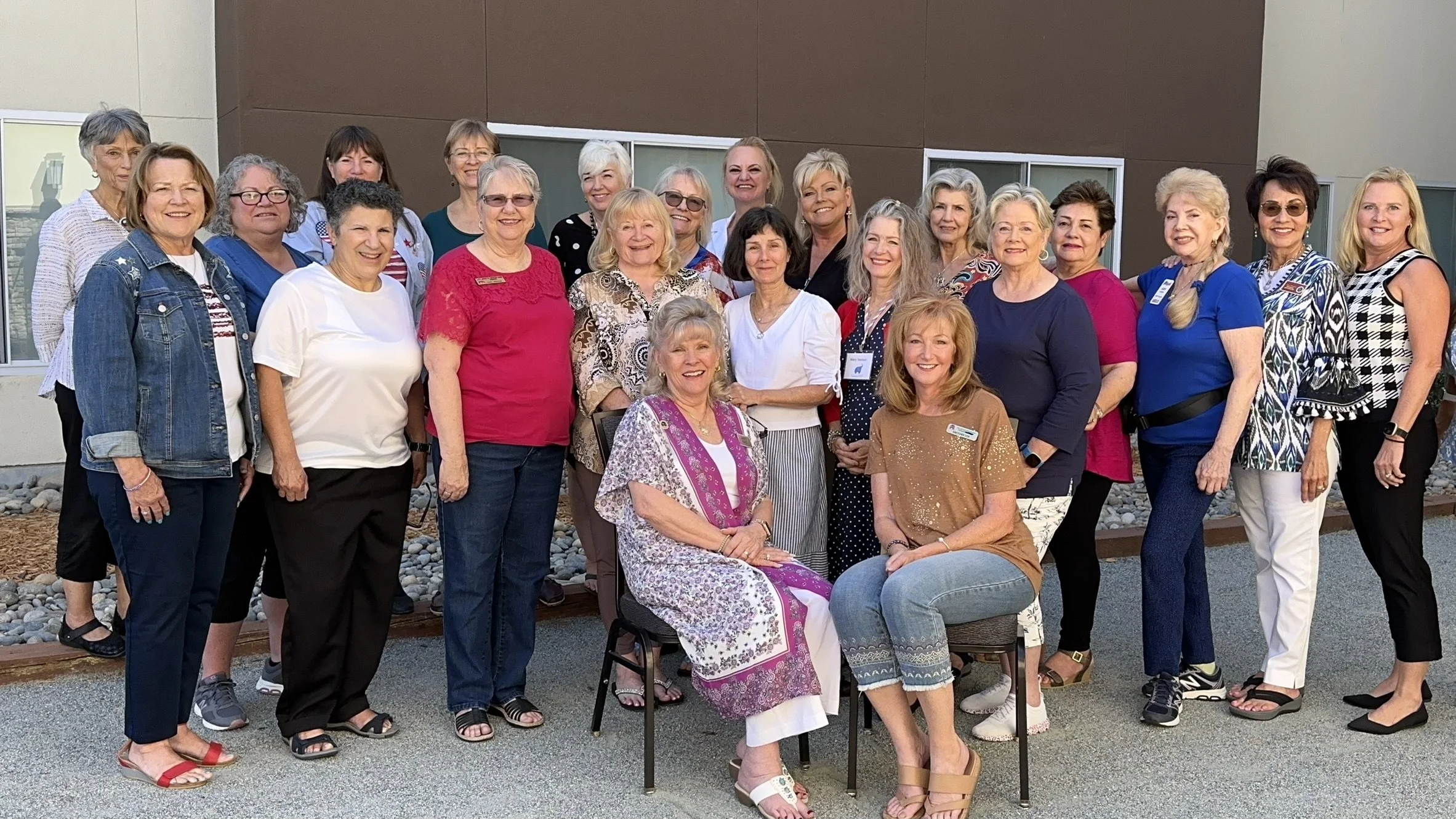 Group of women posing outdoors in front of a building, some wearing patriotic or casual clothing, all smiling.