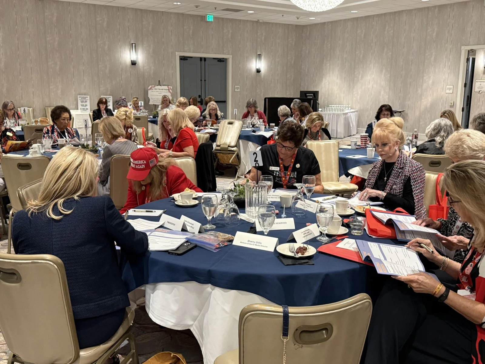 Women seated at round tables in a conference room, engaging with documents and discussing, with some wearing red hats and clothing.