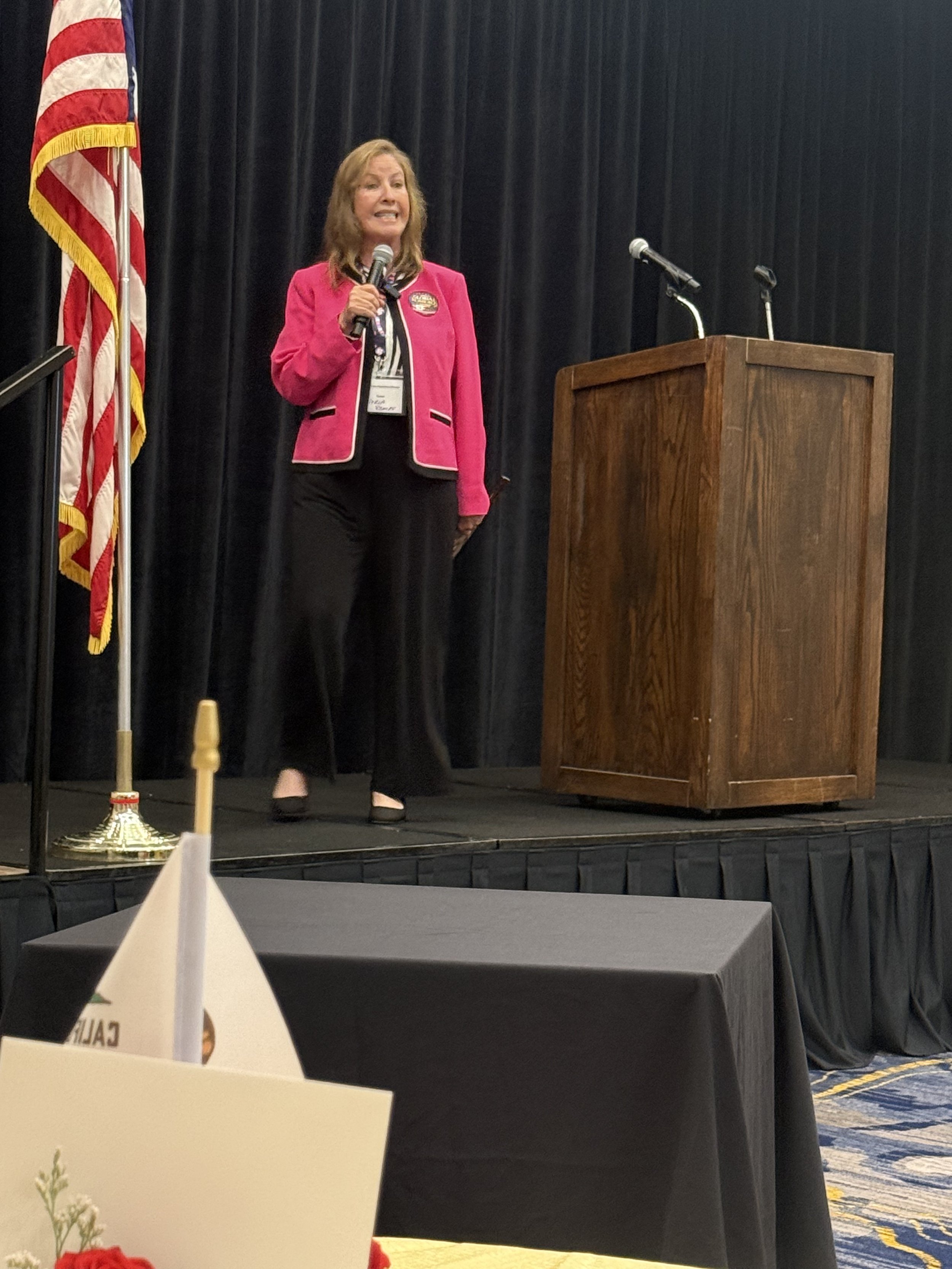 A woman in a pink blazer speaking into a microphone on a stage. Behind her is a black curtain and an American flag. There is a wooden podium on stage.