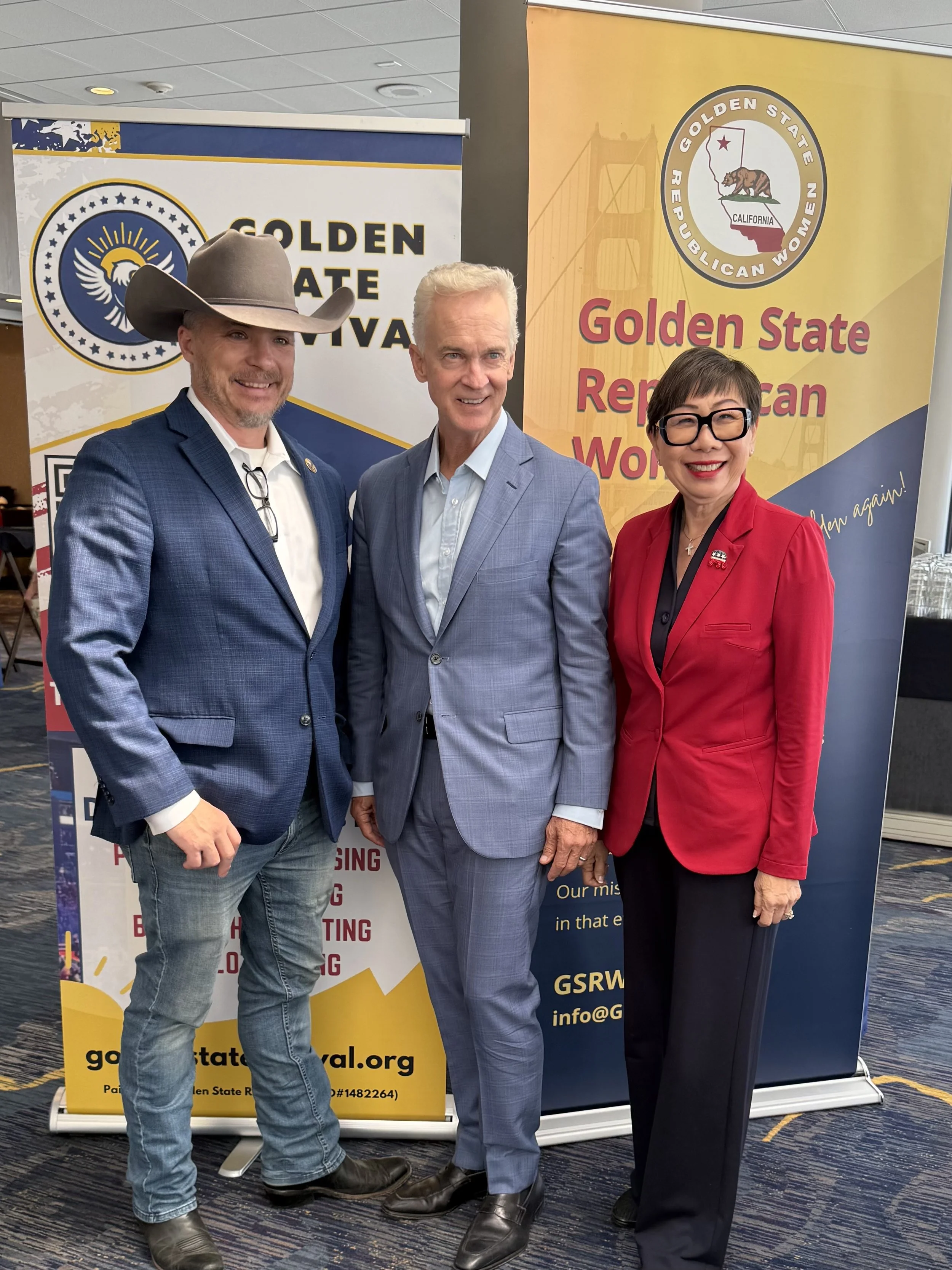 Three people standing in front of banners for the Golden State Republican Women and Golden State Festival. The man on the left is wearing a cowboy hat, a blue blazer, white shirt, and jeans. The man in the middle has white hair, wears a light blue su