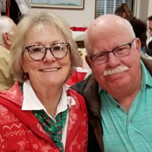 A smiling elderly woman with glasses and a Christmas sweater standing next to an elderly man with glasses and a mustache, in a festive setting.