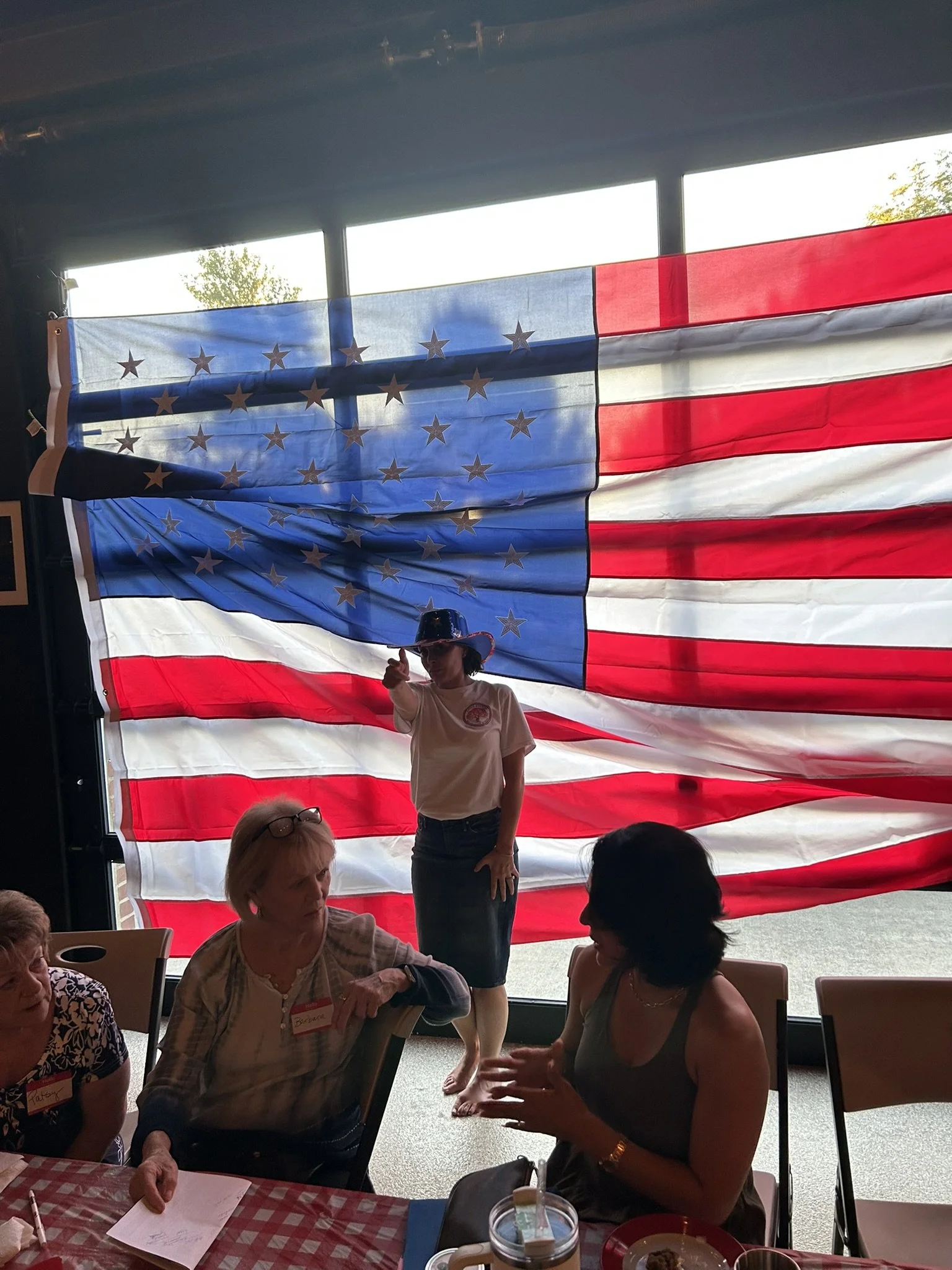 People sitting at a table with a woman standing in front of a large American flag hanging in the background.