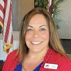 A woman with long brown hair smiling, wearing a red blazer and a name tag, standing next to American flags indoors.