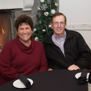 A smiling woman and man sitting at a dining table with black placemats and napkins, in front of a decorated Christmas tree.