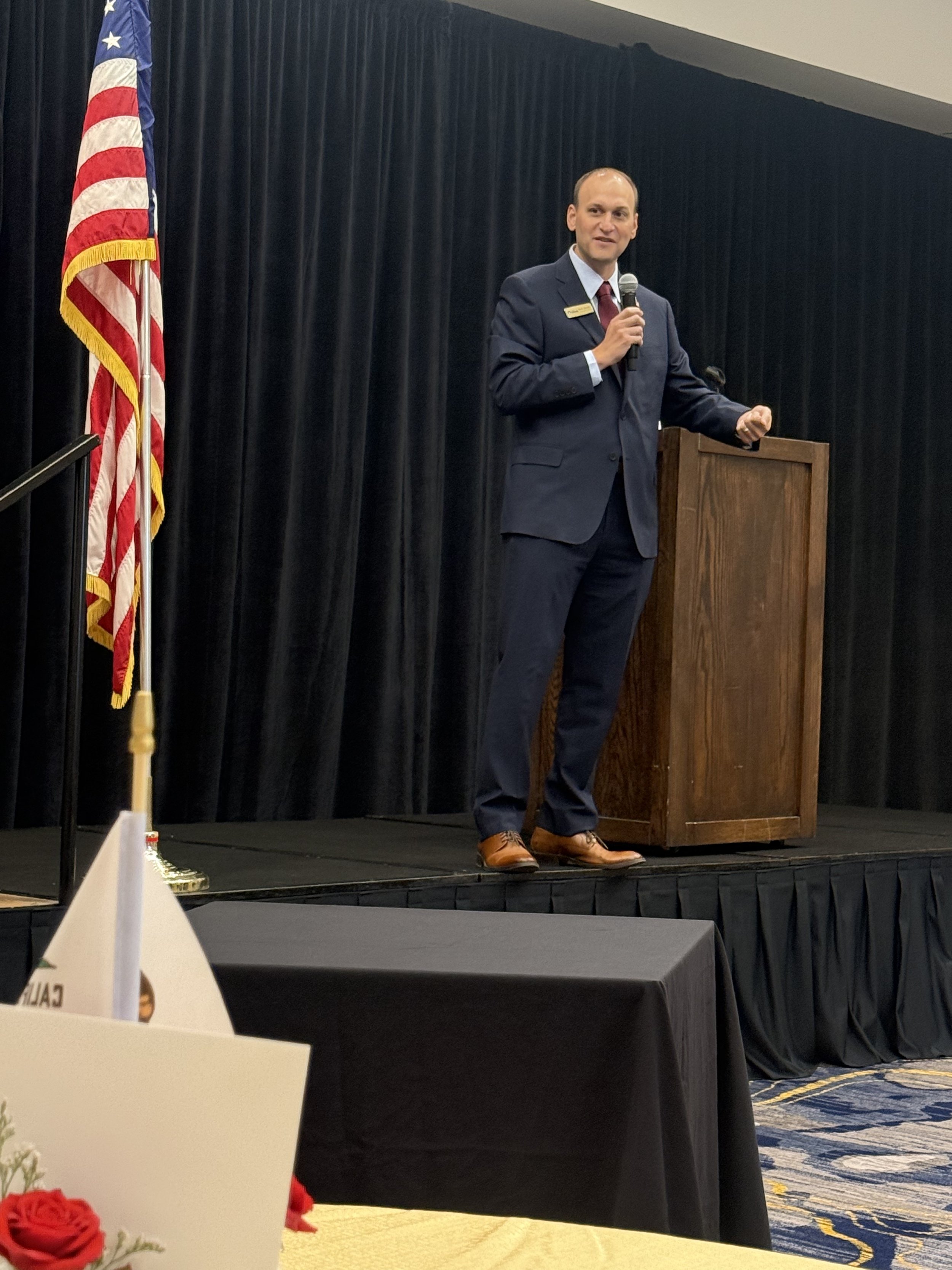 A man in a dark suit and red tie speaking into a microphone at a podium during an event, with an American flag to his left and dark curtains behind him.