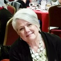 A smiling older woman with short white hair, wearing a black jacket and a white necklace, at a formal event with red tablecloths and chairs in the background.