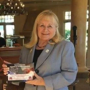 Older woman with blonde hair in a blue blazer holding a flyer, standing in a well-lit indoor space with large windows and ornate decorations.