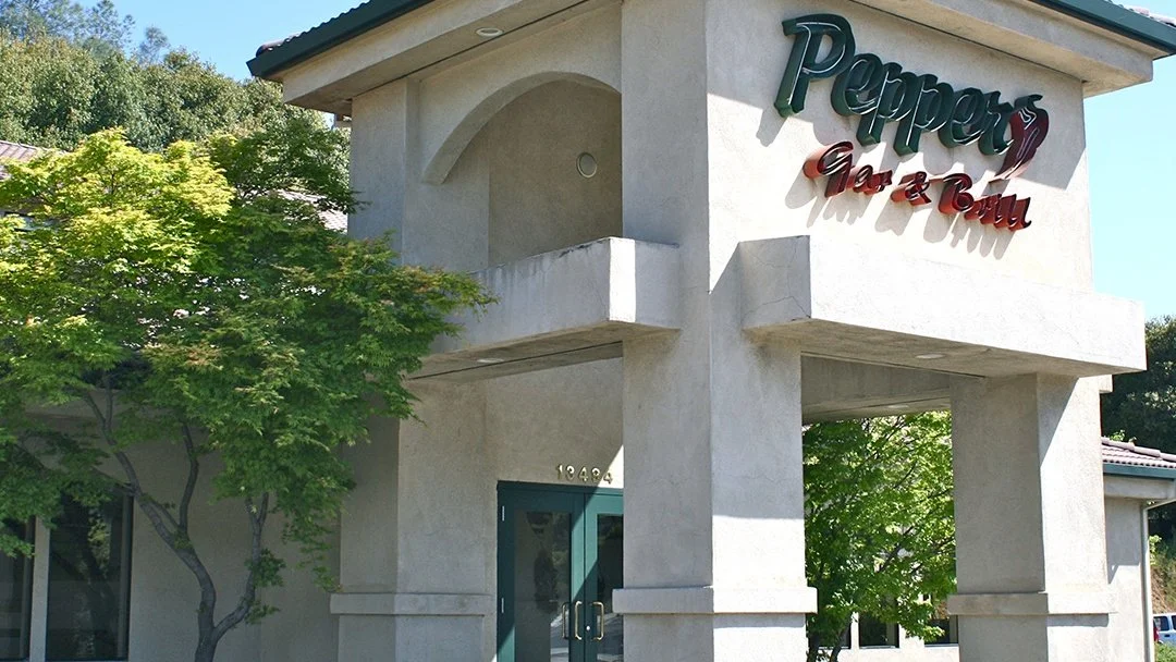 Exterior of a building with a sign that reads "Pepper's Bar & Grill" above the doorway, surrounded by trees and clear skies.