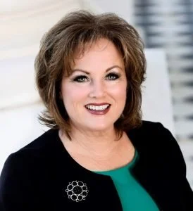 Close-up portrait of a woman with shoulder-length brown hair, smiling, wearing a black blazer with a pin and a teal top, against a blurred background.