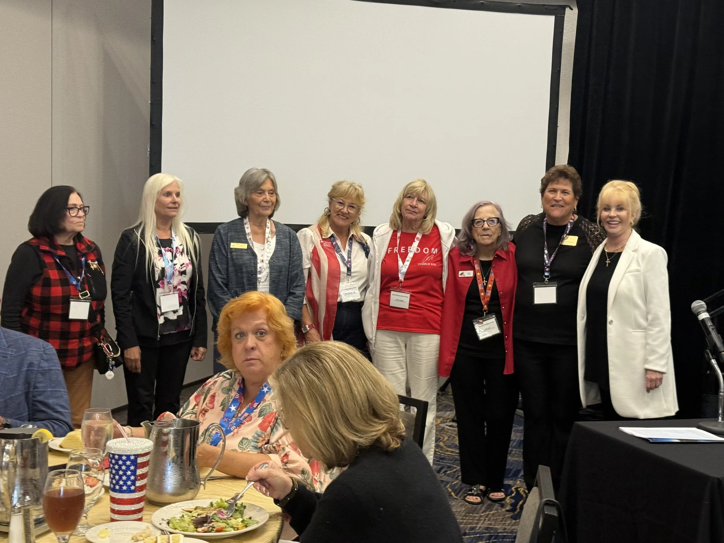 Group of women standing in front of a blank projection screen at a conference or event. They are wearing name tags and some have lanyards. The women are smiling or looking at the camera, and there is someone sitting at a table with plates and drinks 