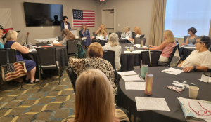 People attending a meeting or seminar in a conference room, seated at tables with papers and drinks, with an American flag on the wall and a large screen at the front.