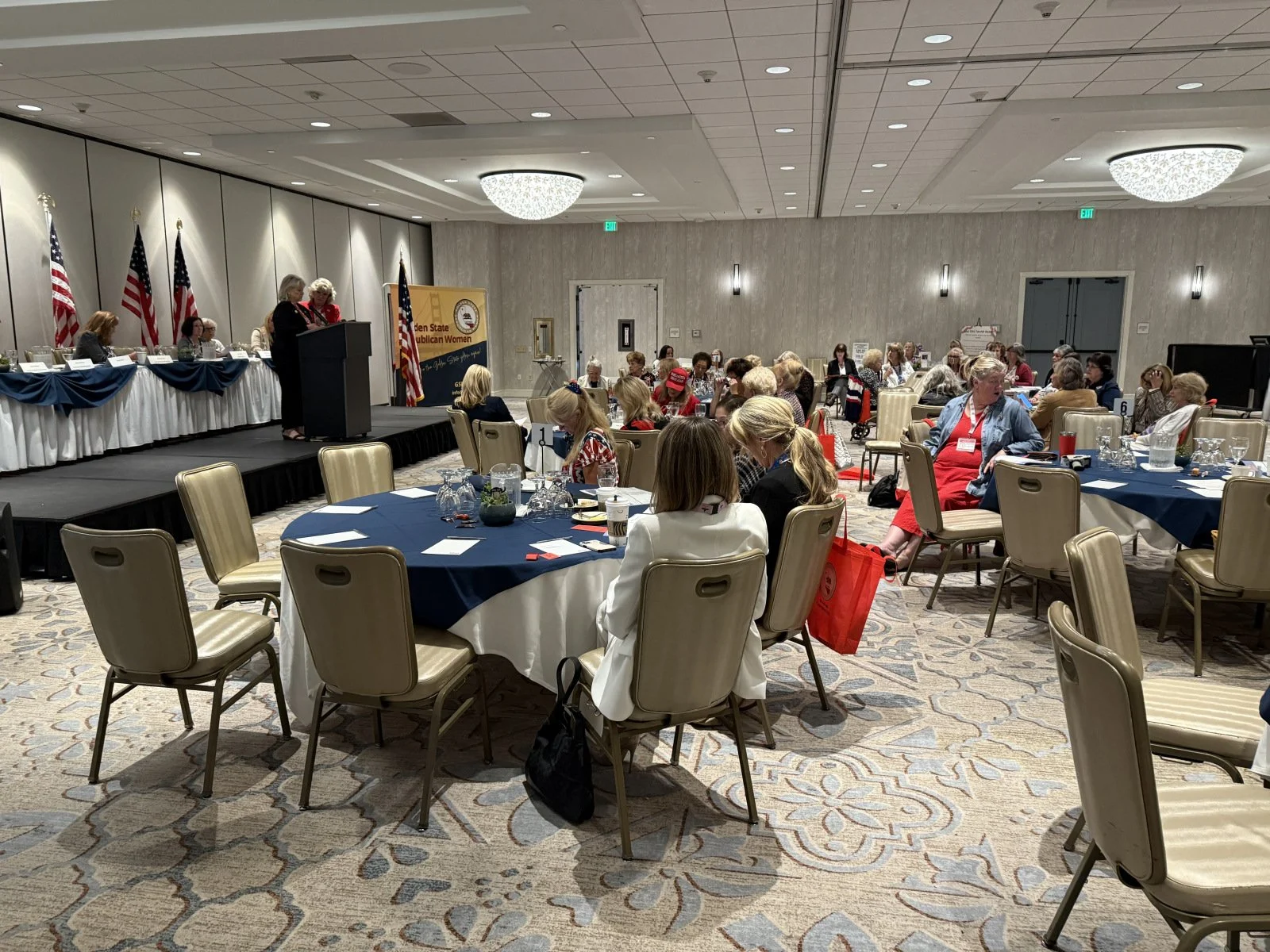 A large conference room filled with attendees sitting at round tables covered with blue tablecloths. Some people are wearing red, white, and blue clothing. There is a stage at the front with a podium, behind which a woman is speaking. American flags 