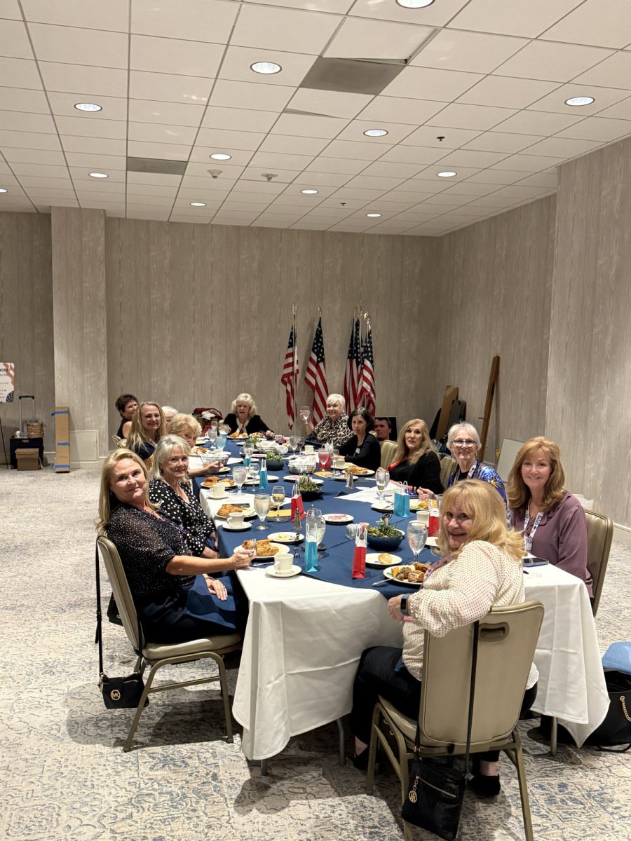 A group of women gathered around a long table during a celebration, with American flags in the background.