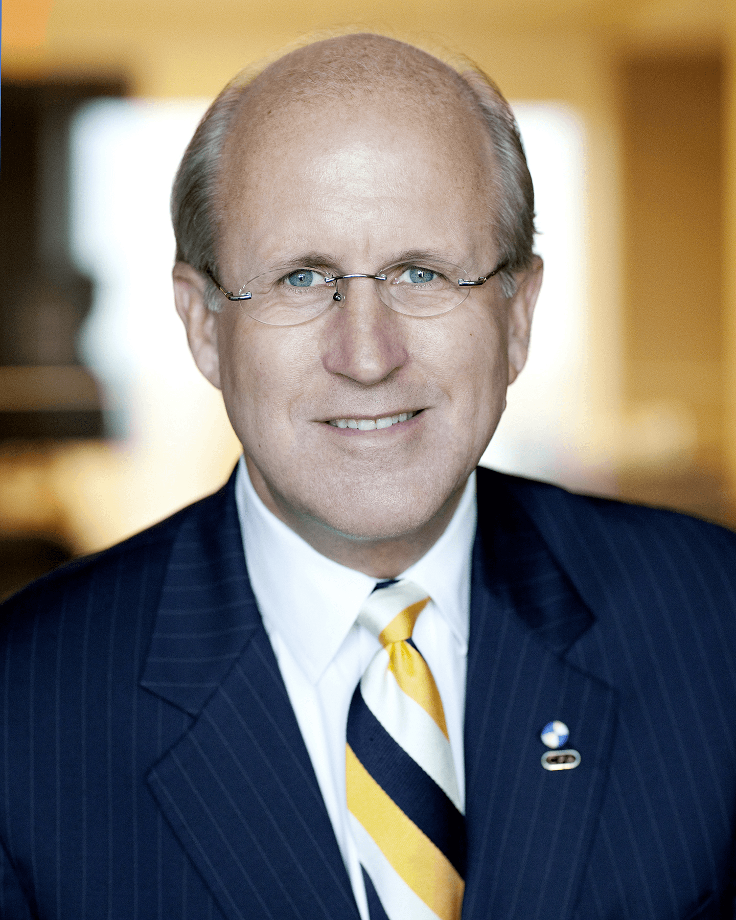 A middle-aged man with blue eyes, wearing glasses, a dark pinstripe suit, a white shirt, and a yellow tie, smiling at the camera.