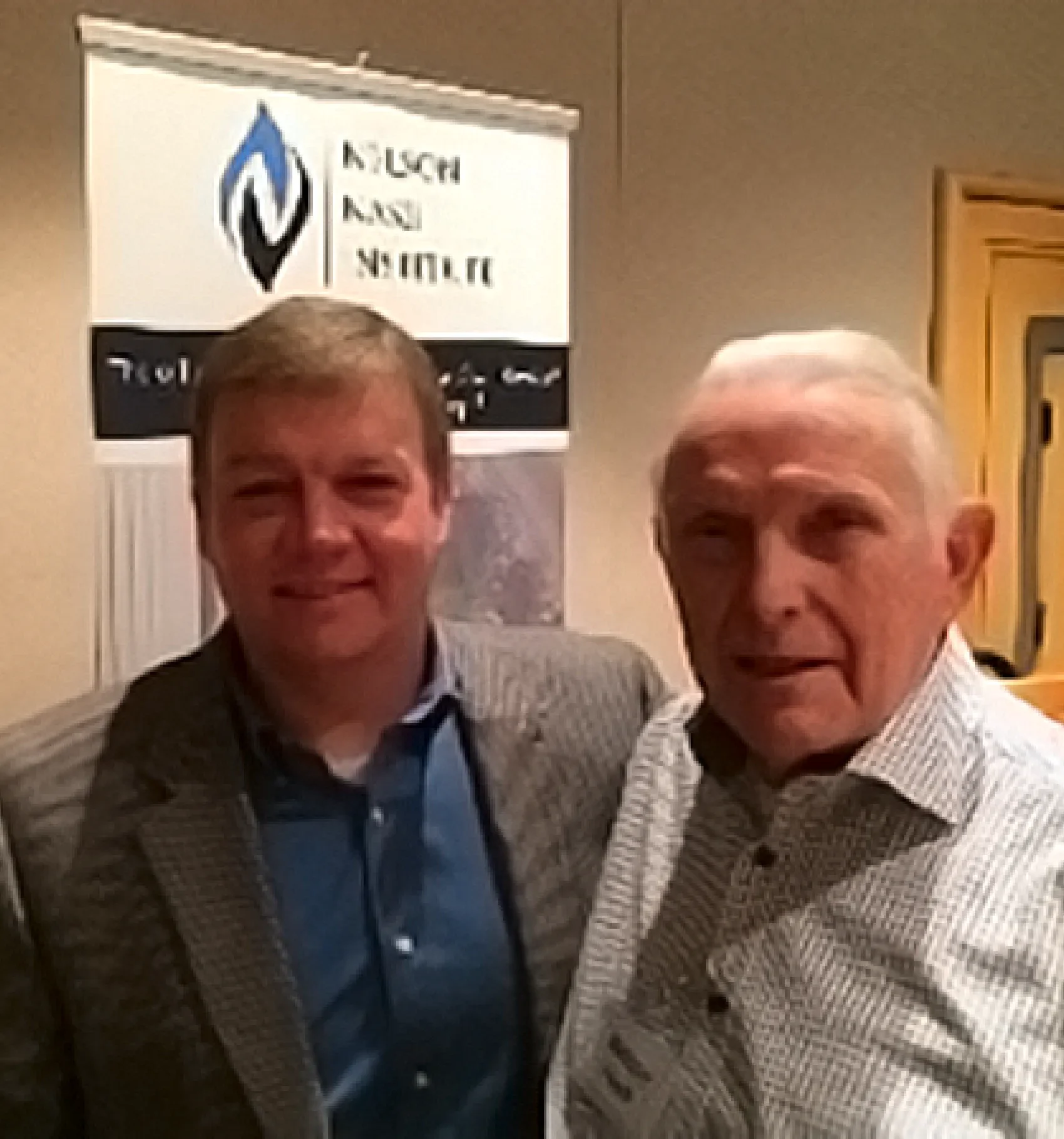 Two men smiling and posing for a photo indoors, with a banner in the background that has a logo and partial text, possibly at an event or meeting.