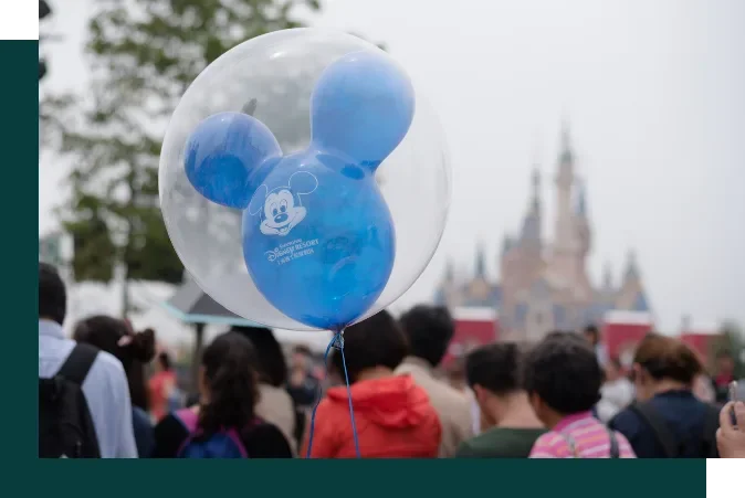 Blue Mickey Mouse-shaped balloon with Disney logo floating above a crowd at Disneyland with the castle in the background.