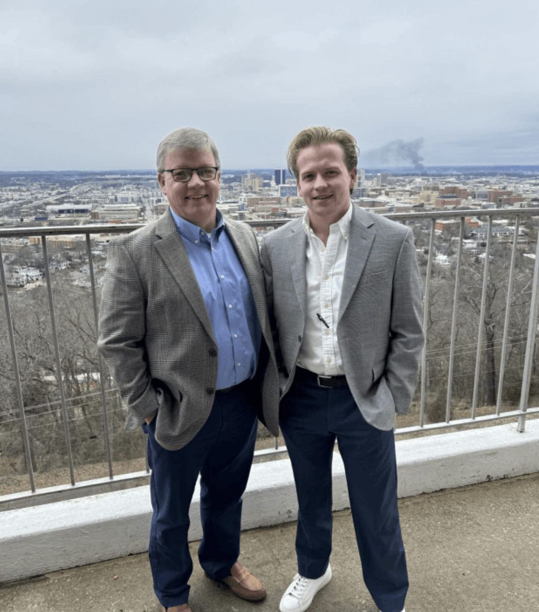 Two men in suits standing outdoors on a balcony overlooking a cityscape with overcast sky.