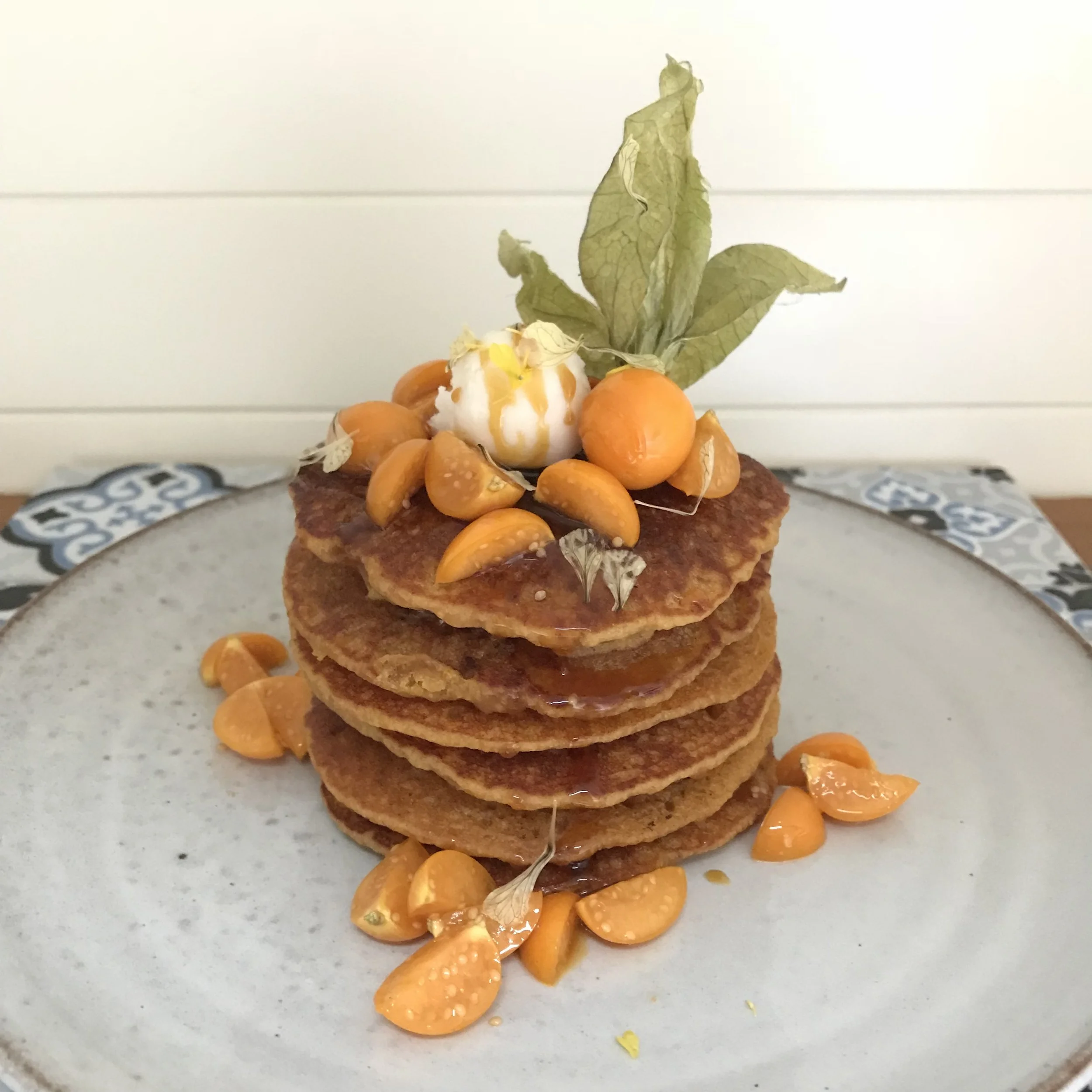 Stack of five caramelized pancakes topped with physalis and a scoop of ice cream, garnished with physalis pieces on the plate and a leaf decor on top.