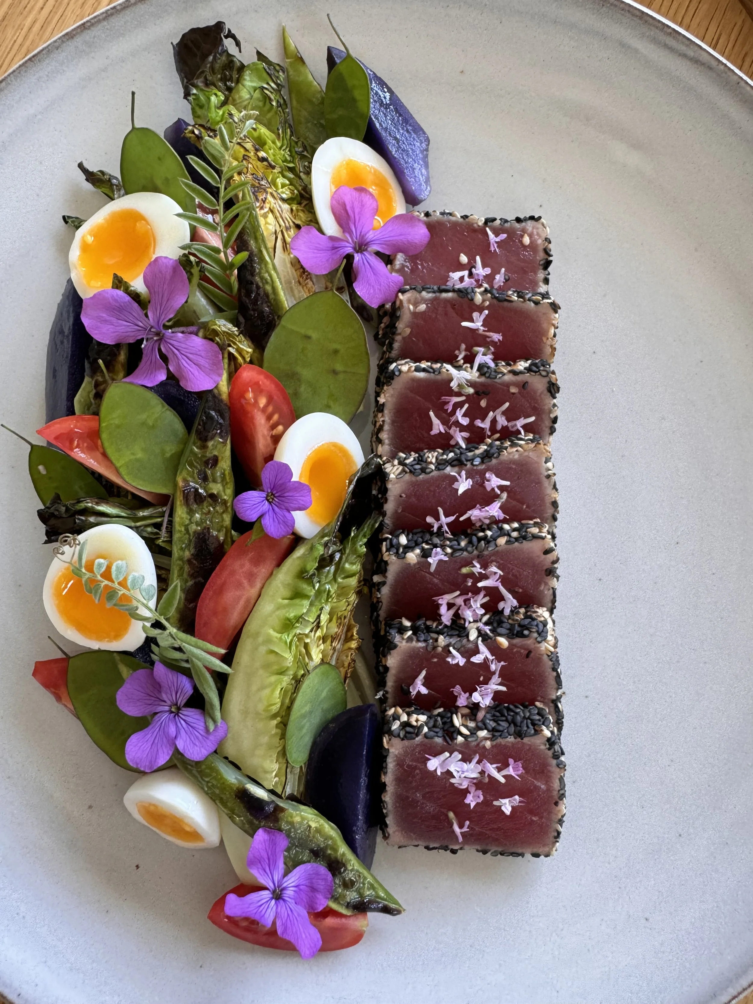 A plate with seared ahi tuna slices sprinkled with pink flower petals, arranged alongside a colorful salad of vegetables including cherry tomato halves, halved soft-boiled eggs, pickles, and edible purple flowers.