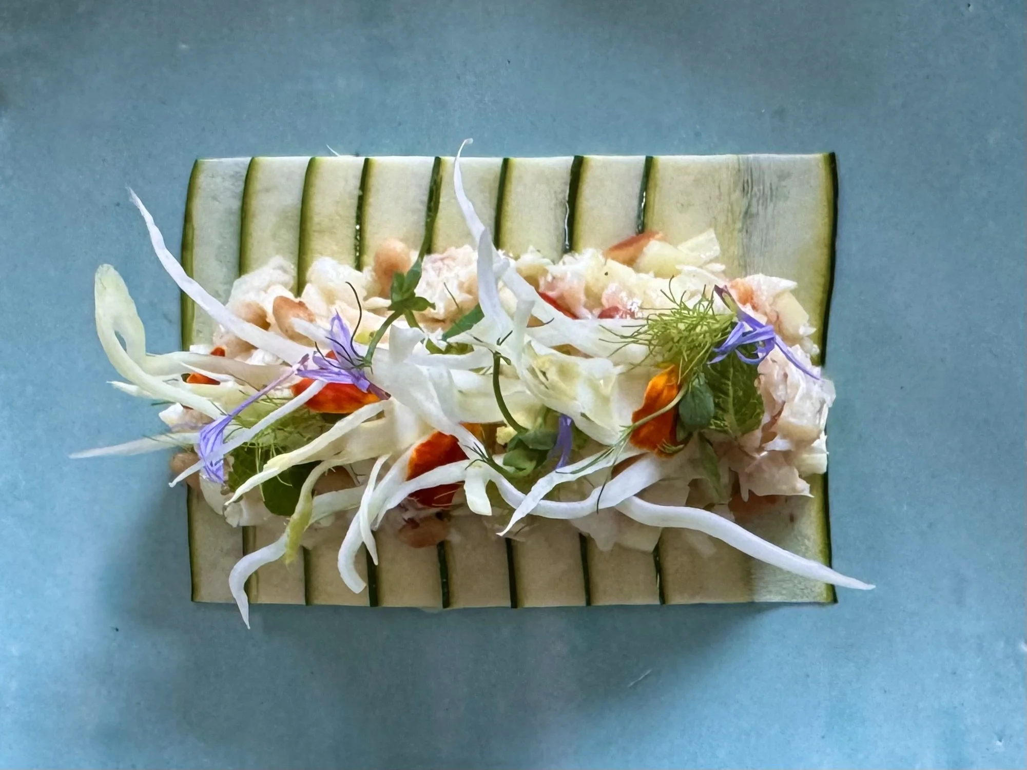 A plated dish of a salad with shredded cabbage, sliced radish, cherry tomatoes, edible flowers, and herbs, served on a rectangular plate with a textured surface.