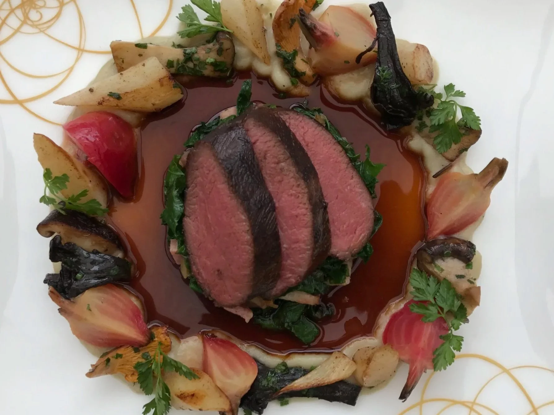 Slices of cooked beef tenderloin topped with a dark sauce, accompanied by roasted vegetables including onions, cherry tomatoes, and herbs, arranged on a white plate garnished with parsley.