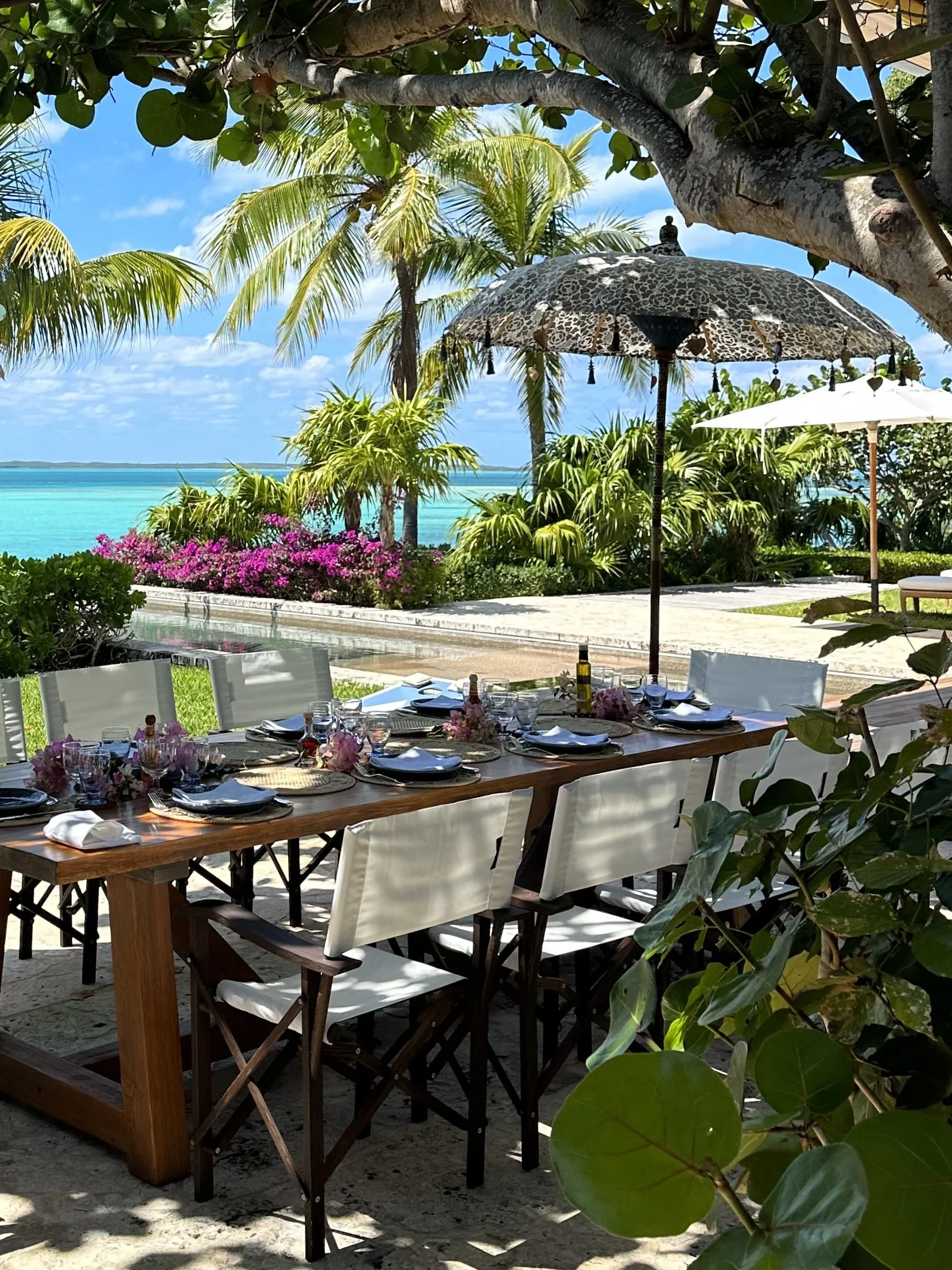 A beautifully set outdoor dining table with white chairs, pink floral centerpieces, and tableware, situated on a sandy area under palm trees overlooking a tropical beach with turquoise water and pink flowers in the background, shaded by an umbrella with a leopard print pattern.