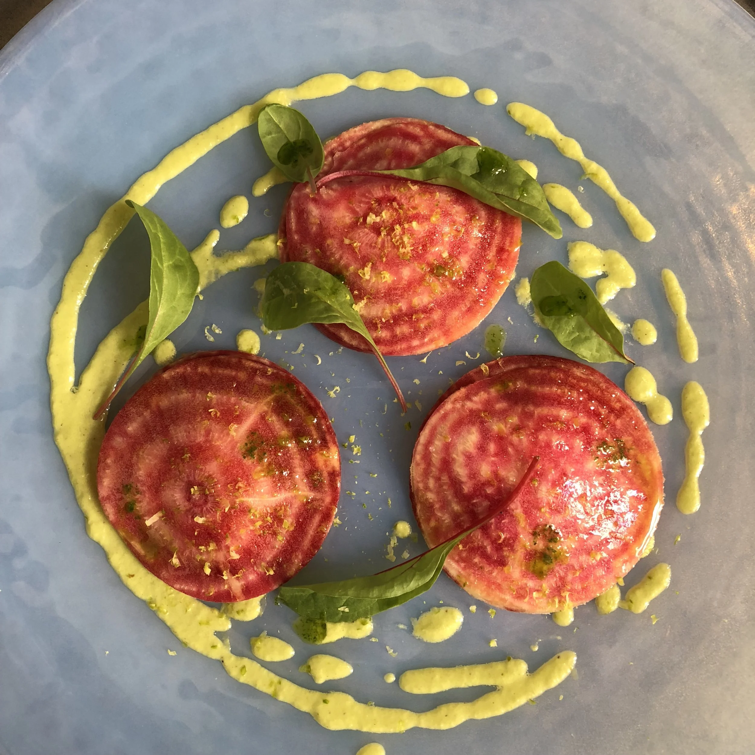 Three halved heirloom tomatoes placed on a light blue plate, garnished with fresh green basil leaves and drizzled with a yellow-green sauce or dressing.