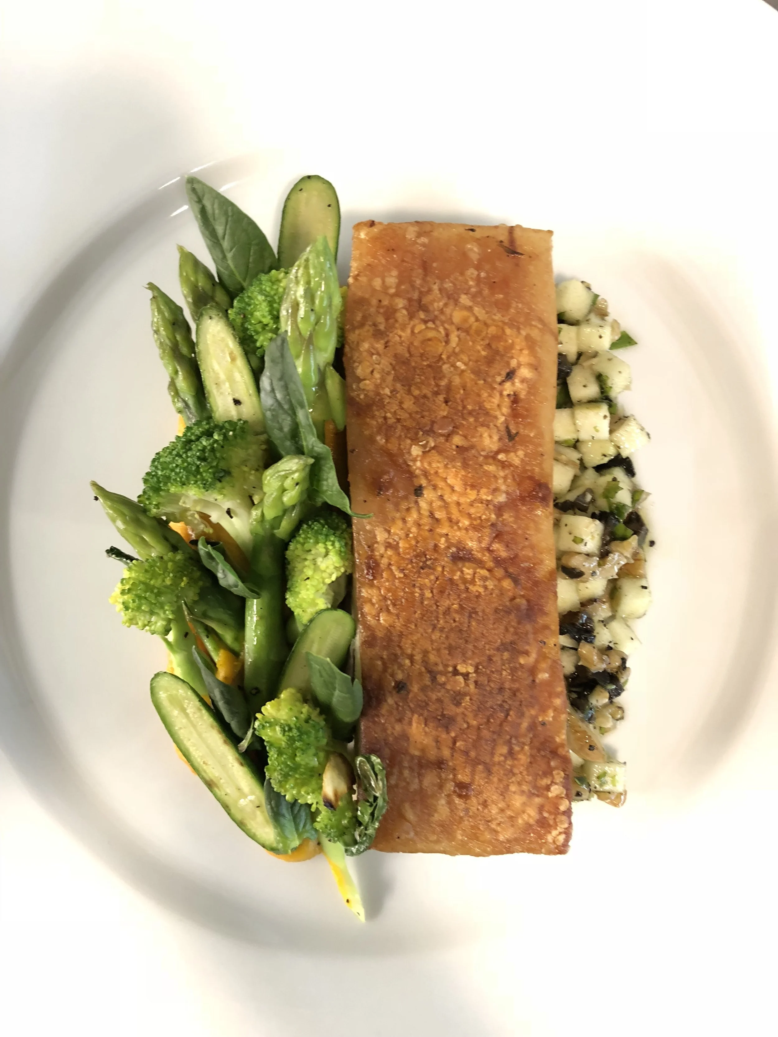 Seared fish fillet served with roasted vegetables including zucchini, asparagus, broccoli, and spinach, accompanied by a mixed grain salad.