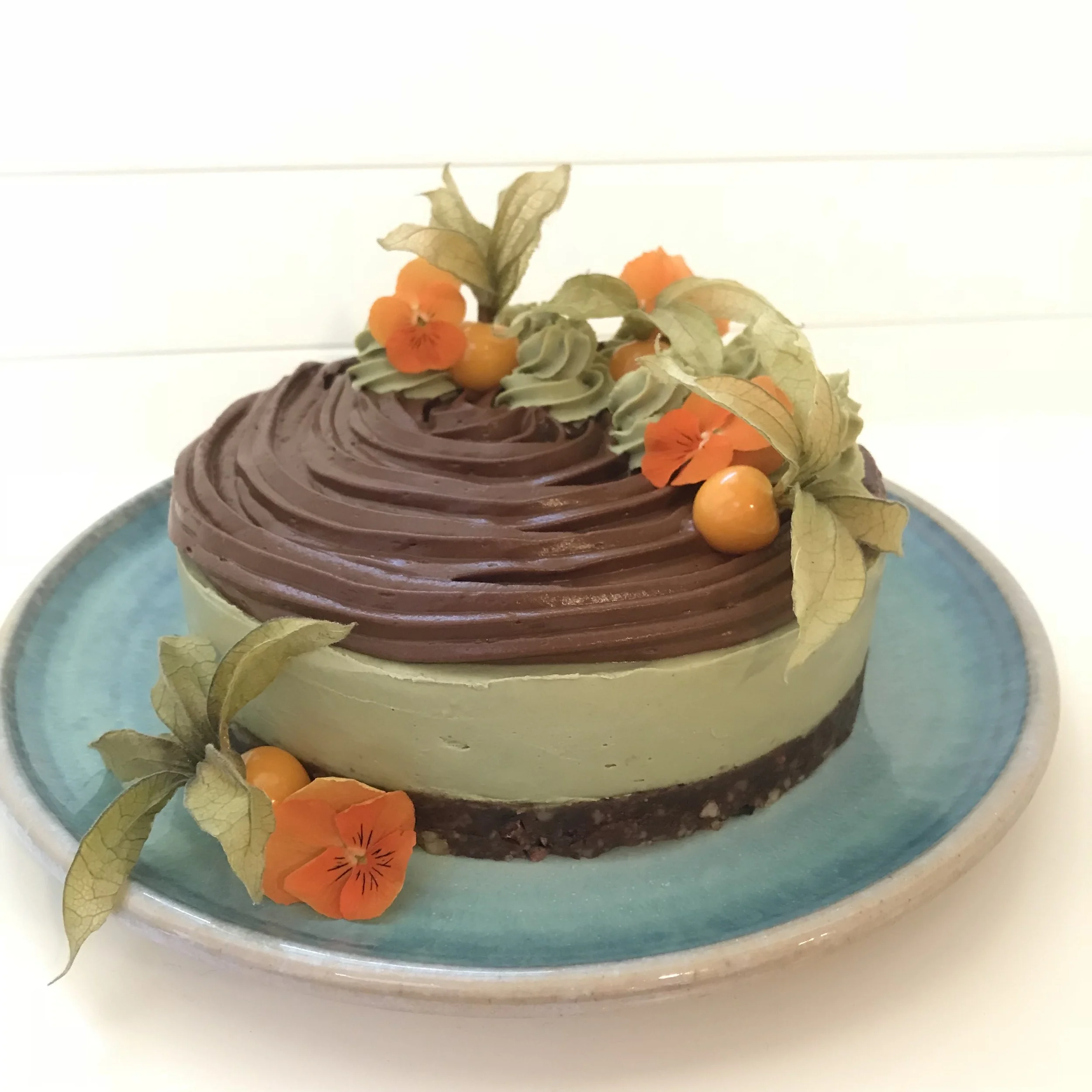 A layered dessert cake with green and brown layers, decorated with orange berries, orange flowers, and dried leaves on a blue plate.