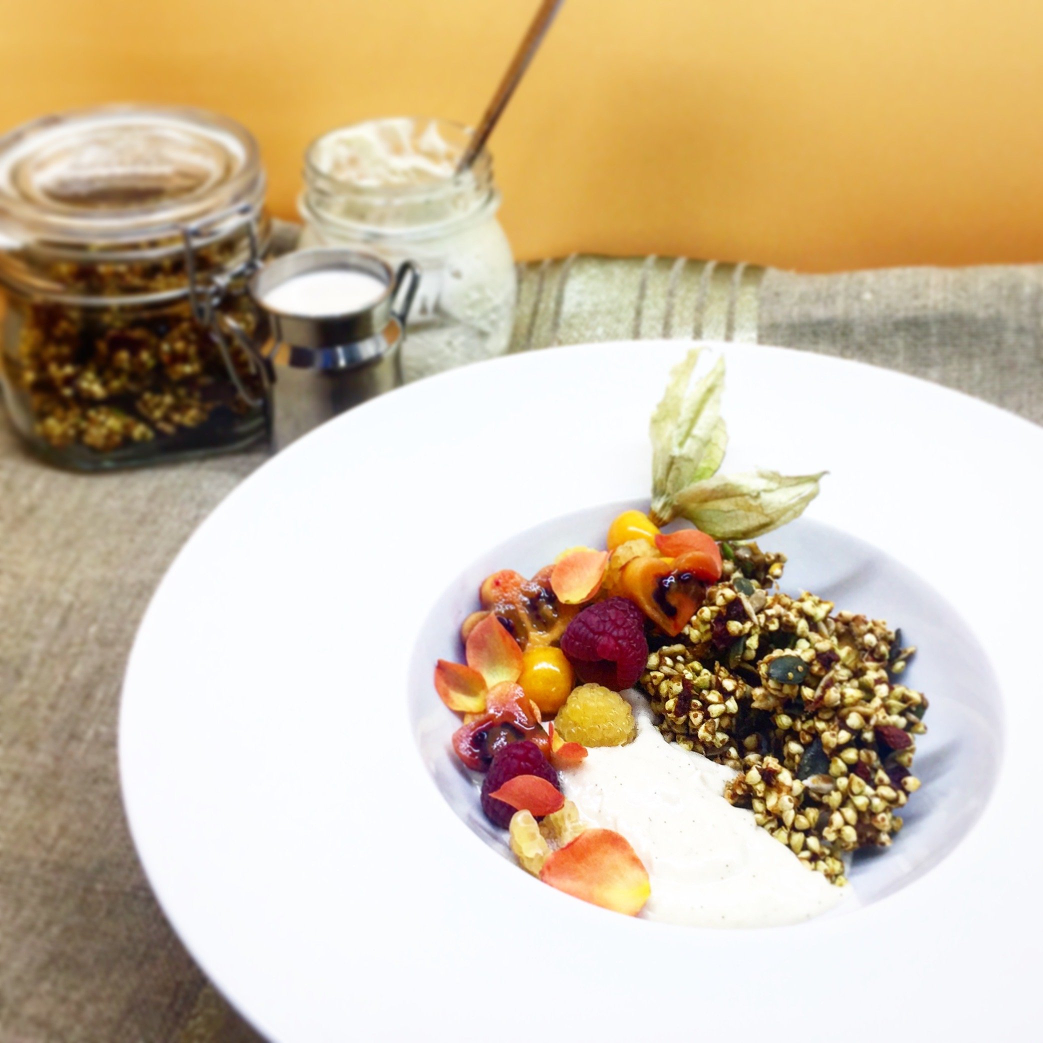 Granola with mixed berries, sliced banana, and a sprig of dried leaves on a white plate, with jars of spices, honey, and yogurt in the background.