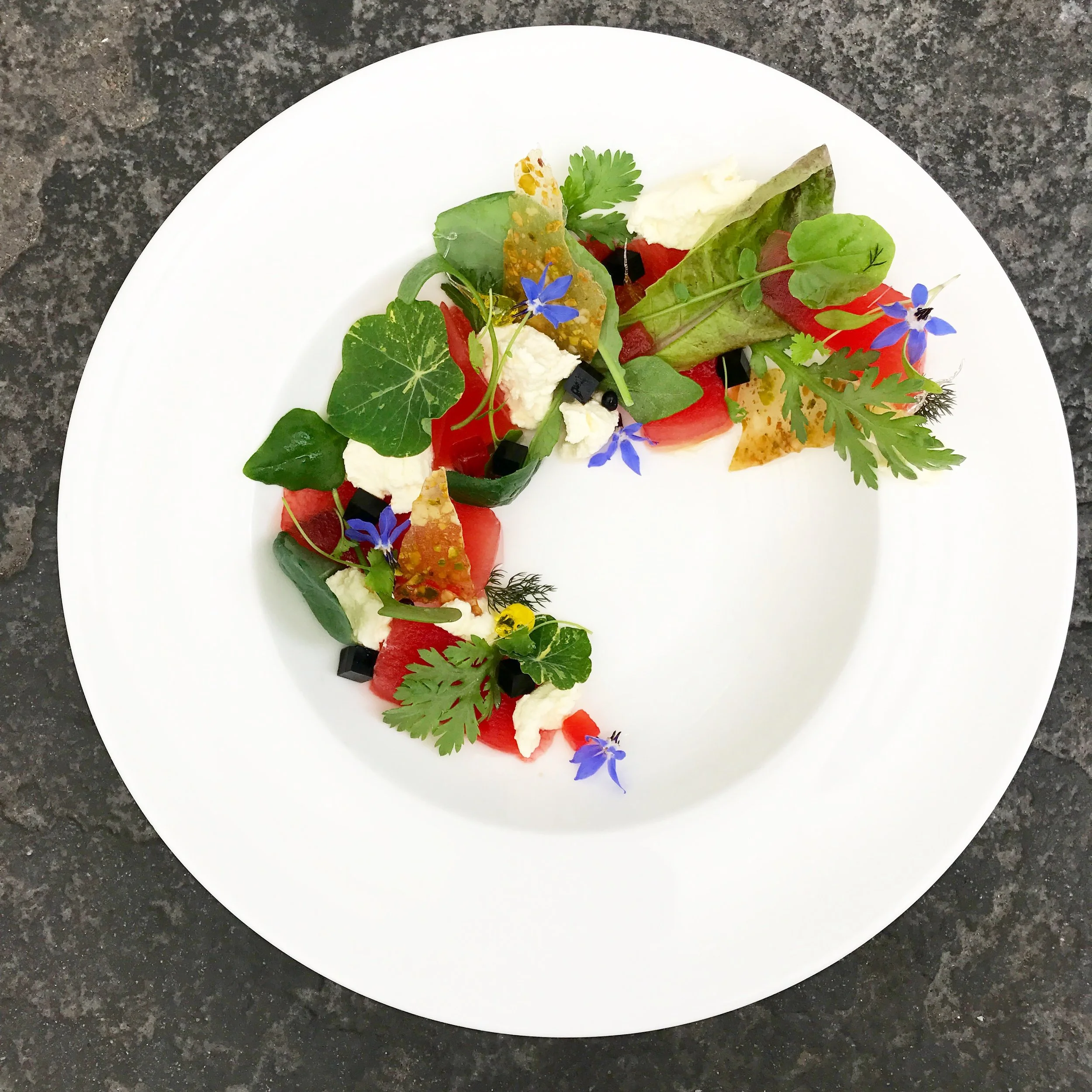 Artistic plate of fresh salad with assorted leafy greens, edible flowers, diced vegetables, and cheese garnished with herbs.