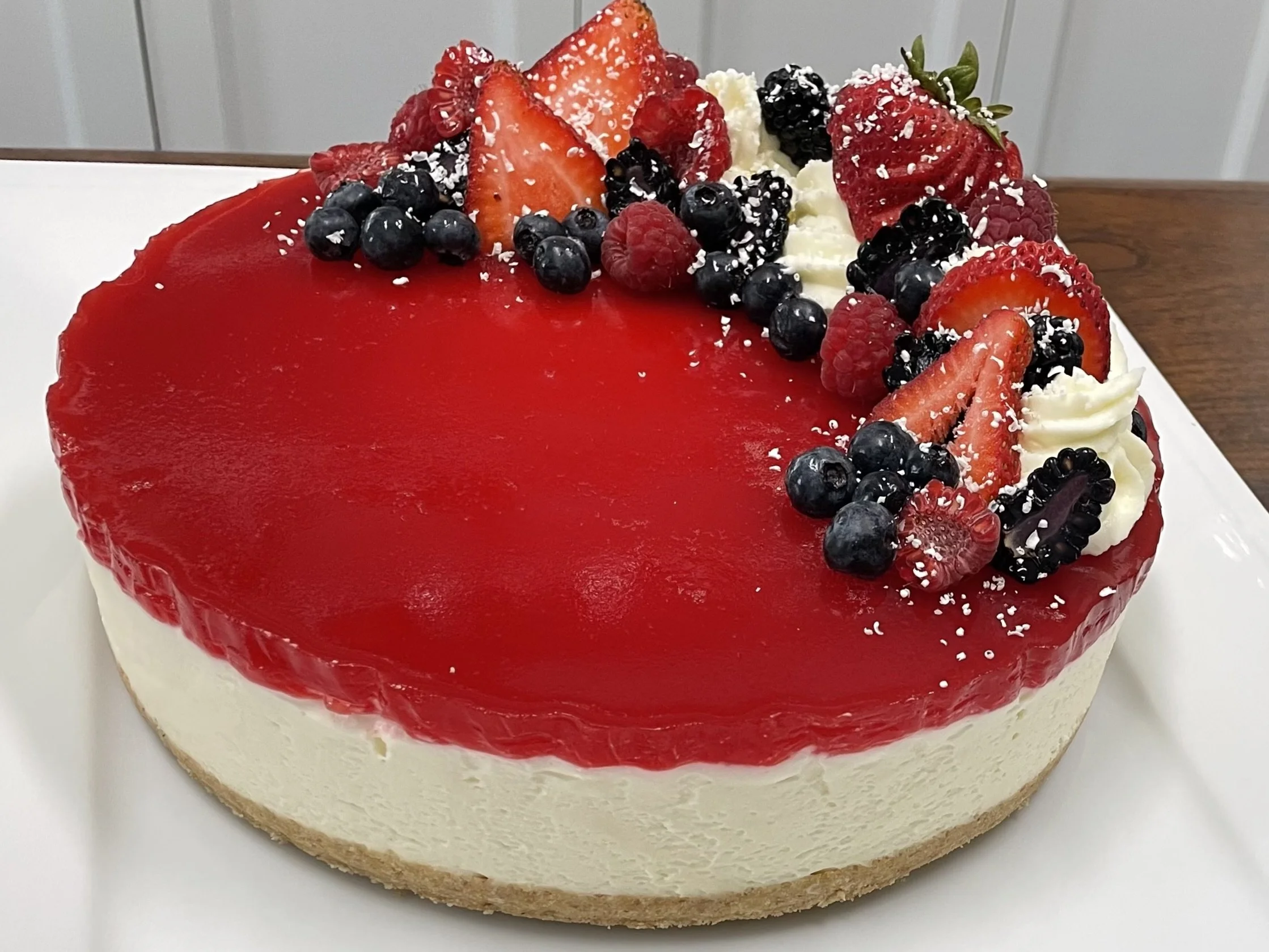 Cheesecake topped with a red glaze and fresh berries, including strawberries, blueberries, and raspberries, garnished with white sprinkles.
