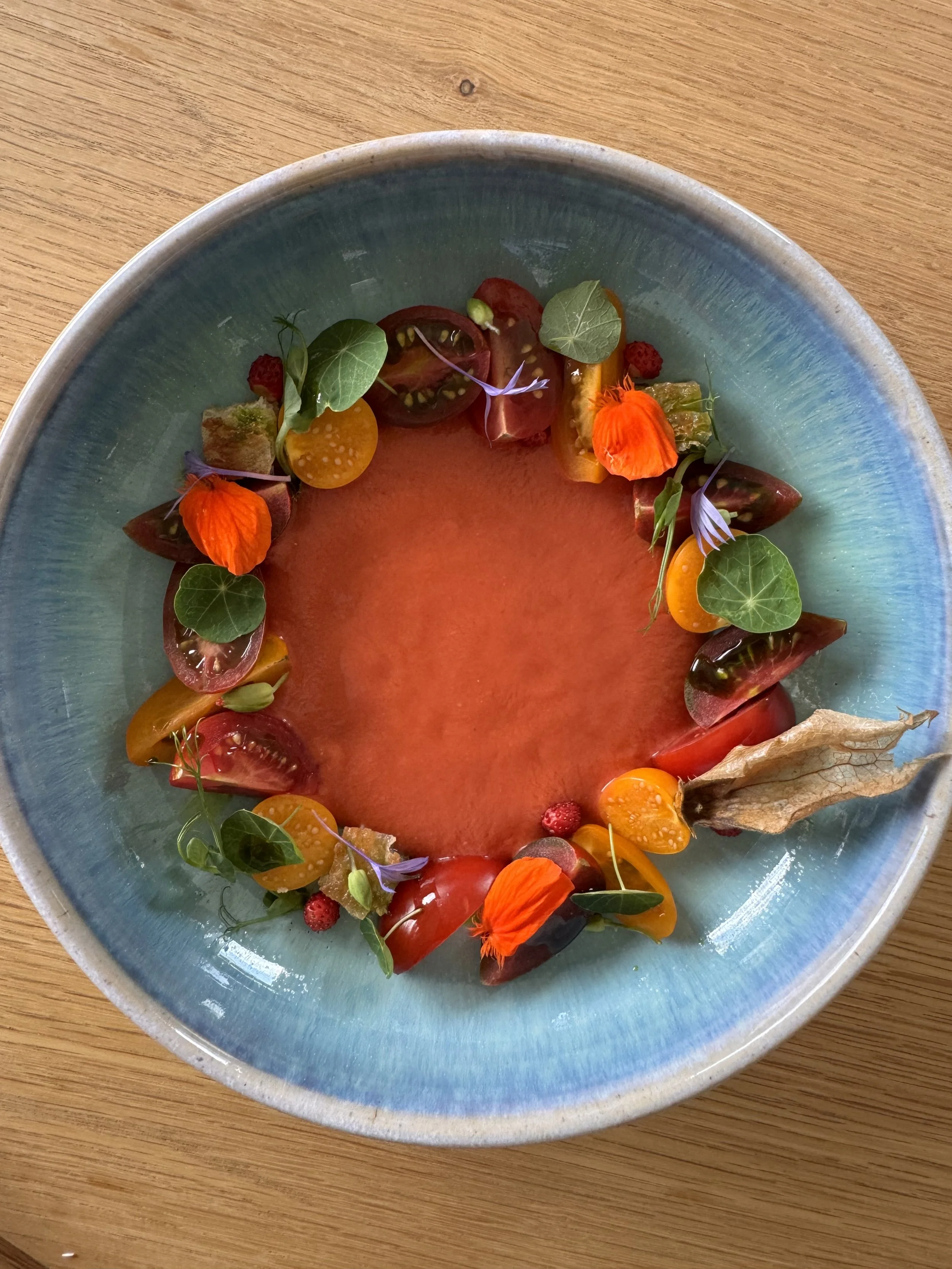 A bowl of watermelon with various fresh tomatoes, edible flowers, and greens arranged around the rim, creating a colorful presentation.