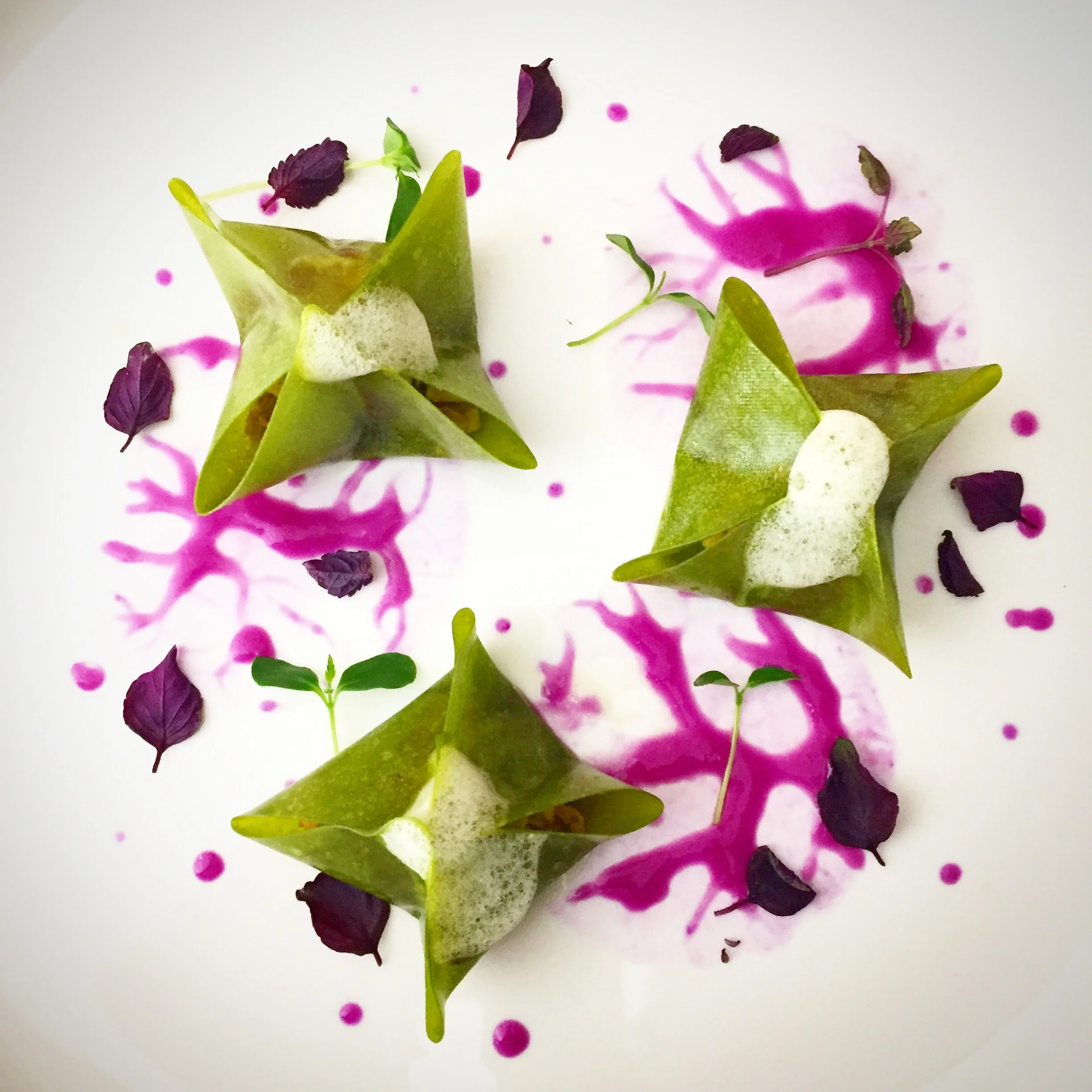 A plated dish with three green, star-shaped, edible confections topped with foamy white sauce, garnished with purple edible flowers and tiny green herbs on a white plate with pink sauce splashes.