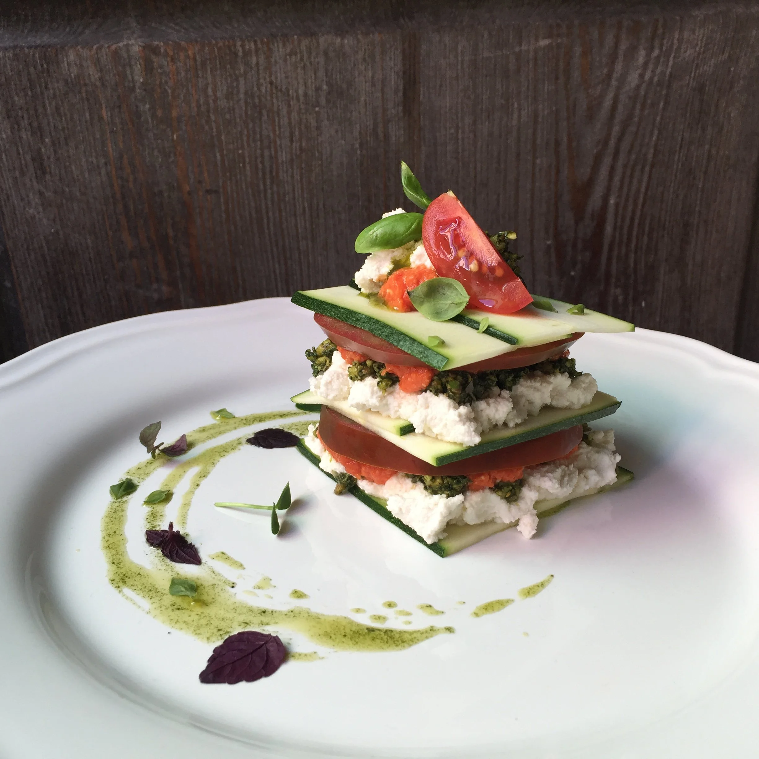 Stacked vegetable terrine with sliced tomatoes, cucumber, and herbs, garnished with microgreens and drizzled with green sauce on a white plate.