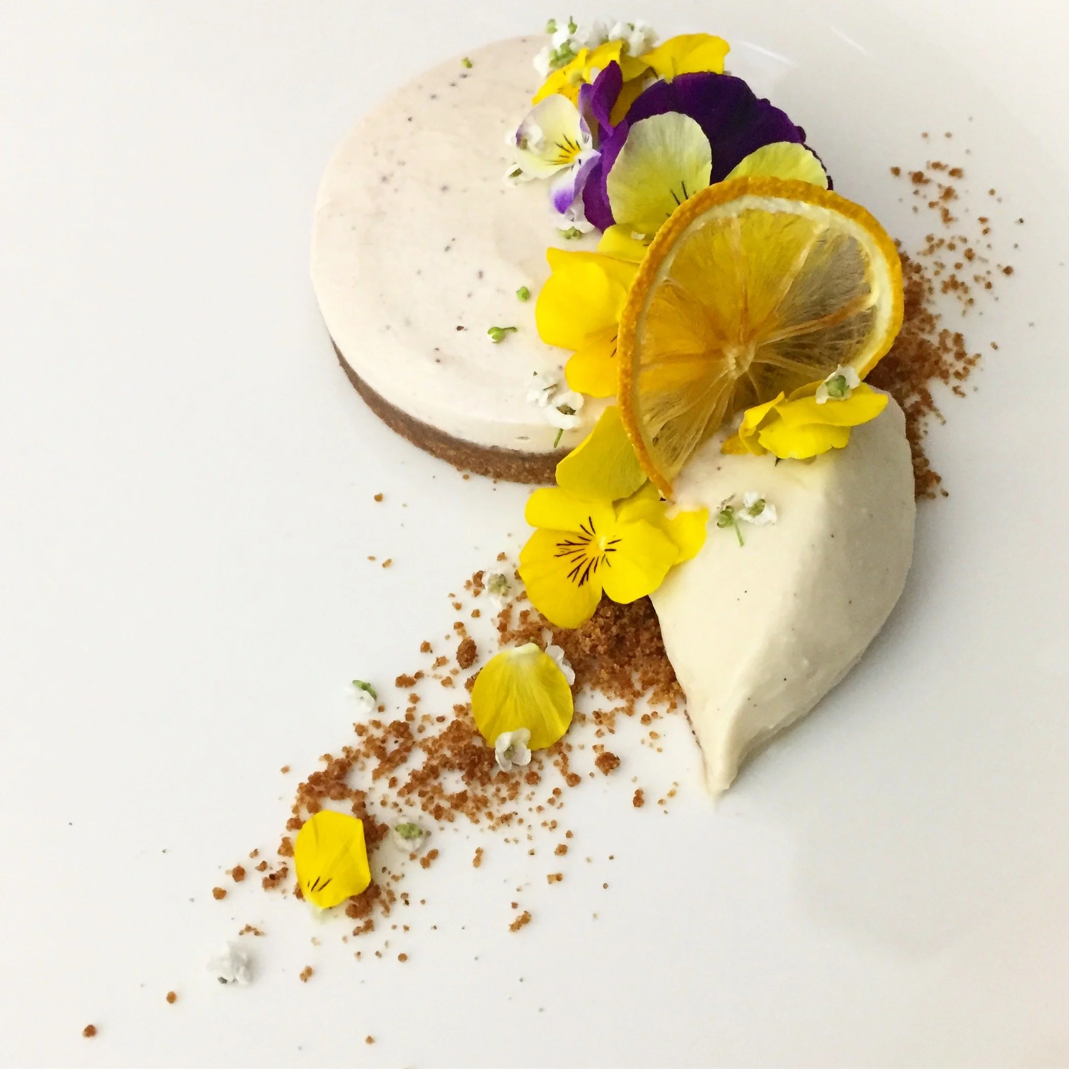 Decorative plated dessert with two mousse-like scoops, dried lemon slices, edible flowers, crushed graham crackers, and small white and yellow garnishes on a white plate.