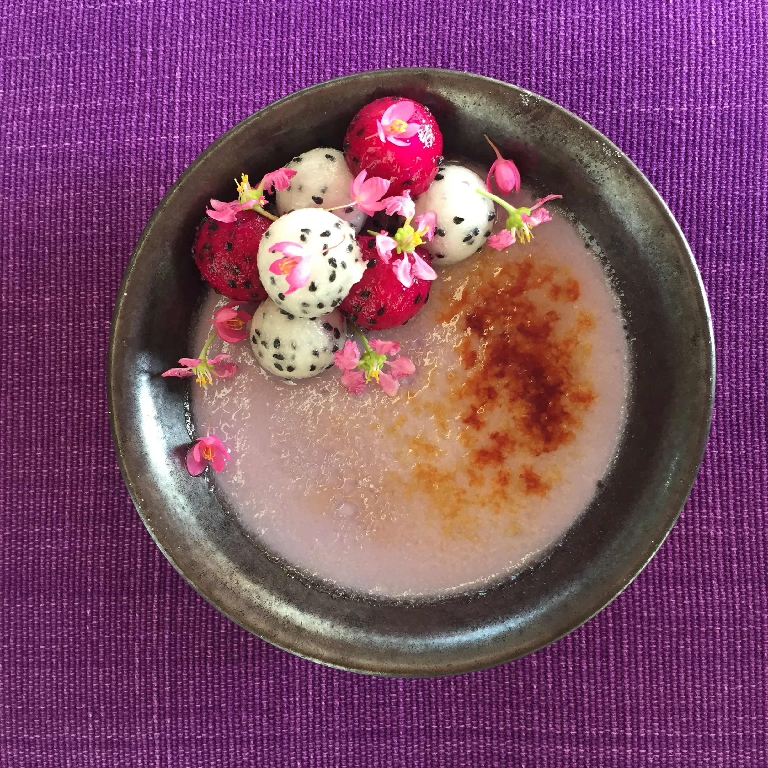 A black bowl with colorful dragon fruit and tongue roses, set on a purple textured tablecloth.
