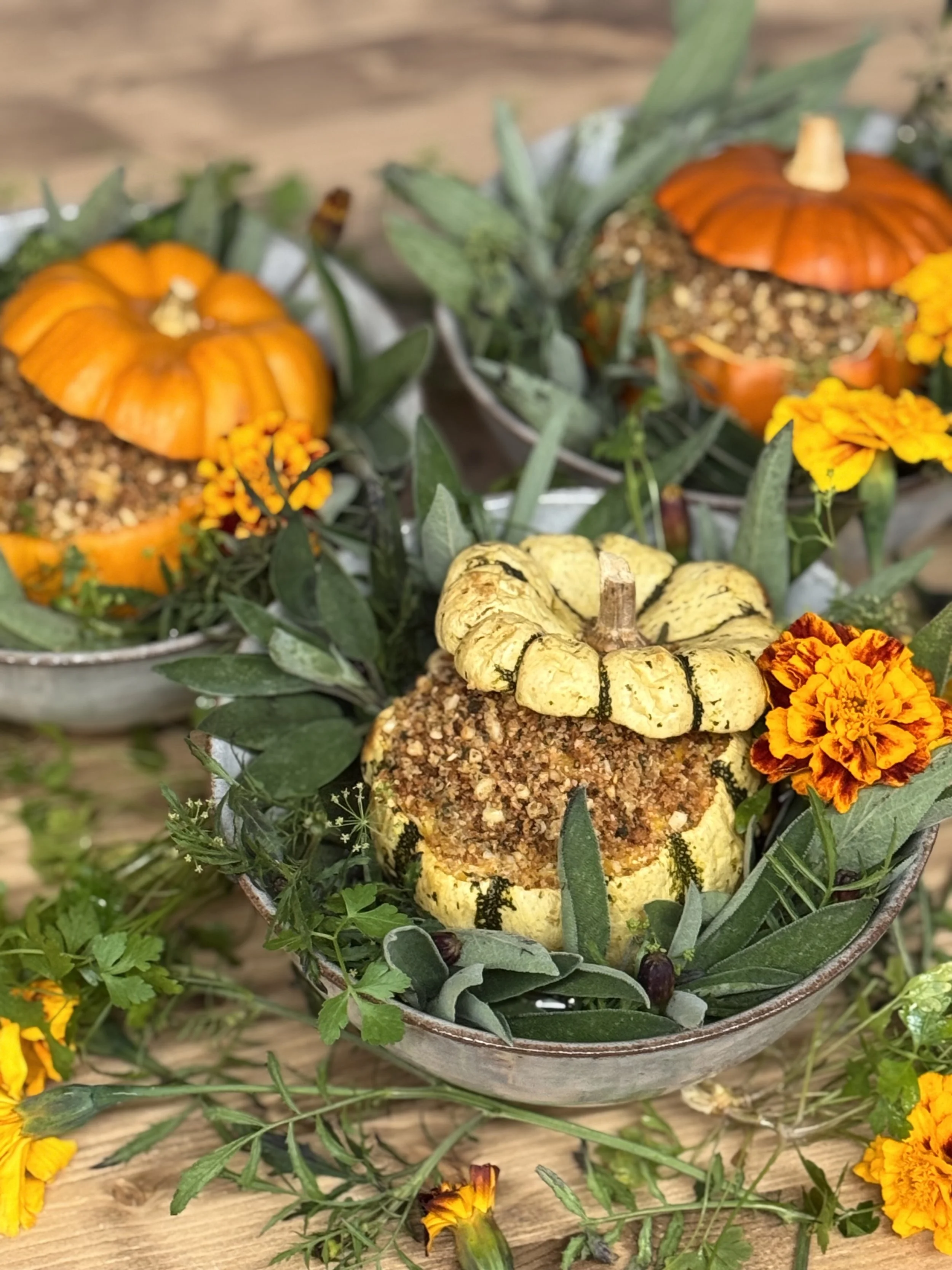 Decorative fall display featuring small pumpkins, arranged in bowls surrounded by greenery and vibrant orange and yellow marigold flowers, on a wooden surface.