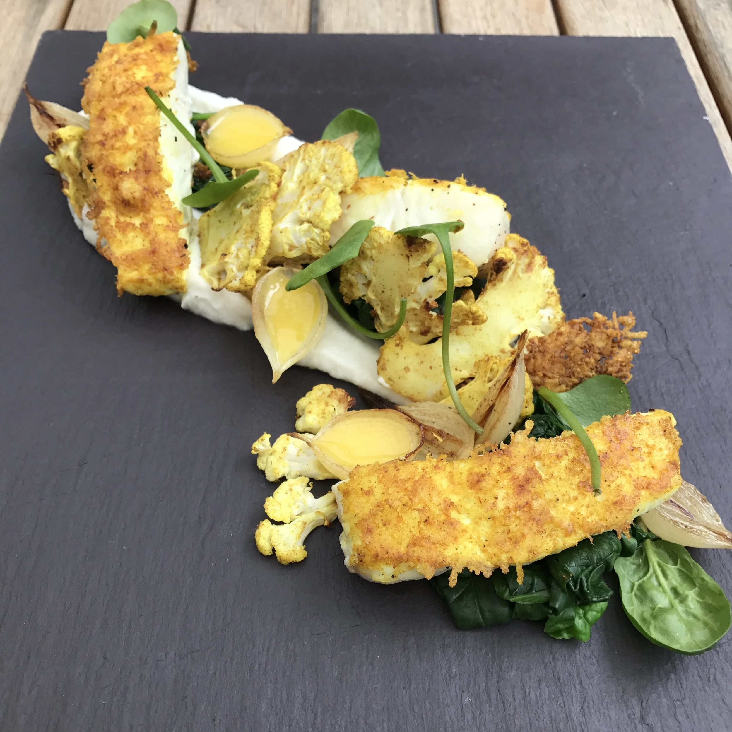 Breaded fried fish fillet on a bed of greens with cauliflower, garlic, herbs, and a light sauce, plated on a black slate serving platter.