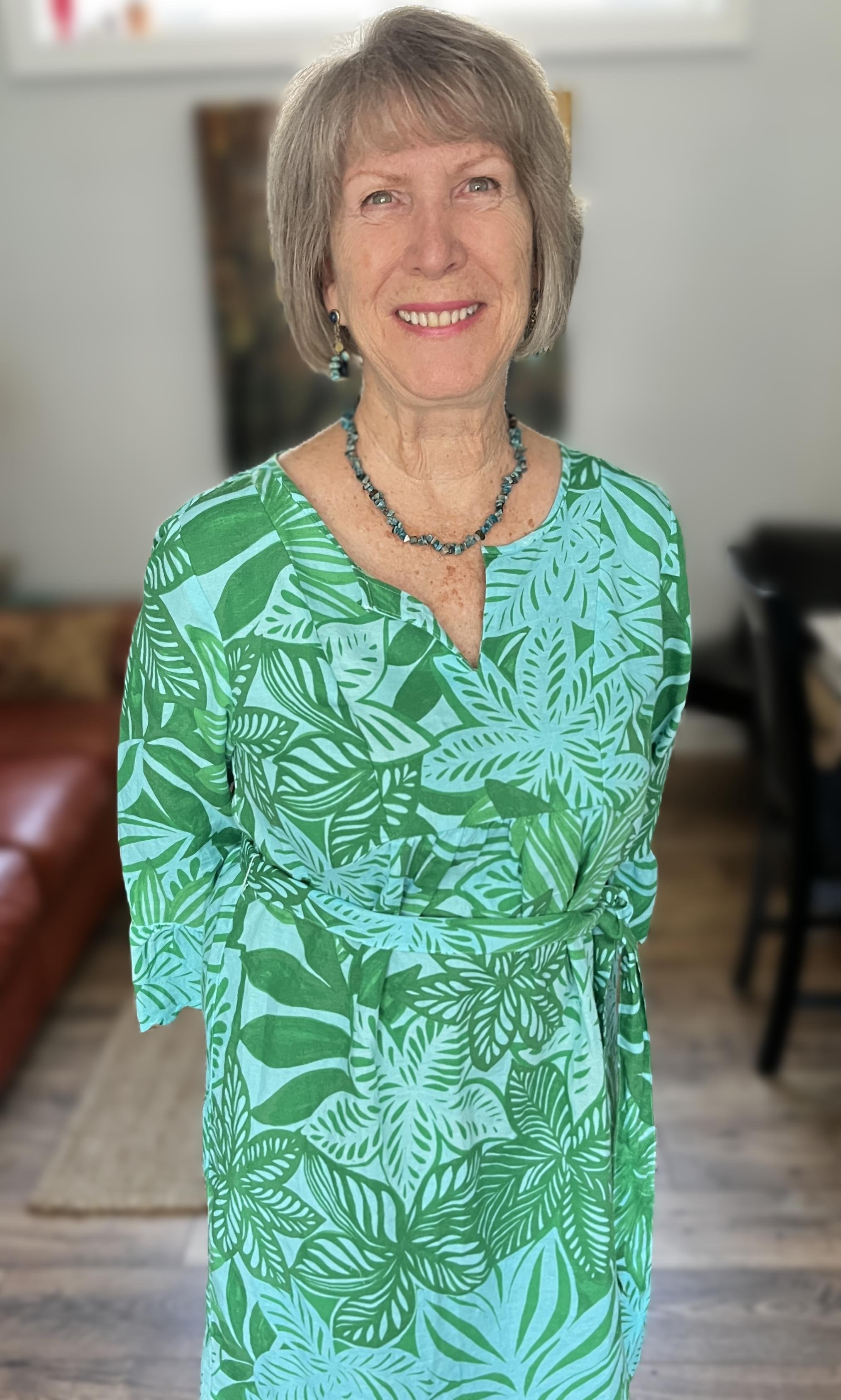 Smiling elderly woman with short gray hair, wearing a green leafy patterned dress, a matching necklace, and earrings, standing in a home with a sofa and a dining area visible in the background.