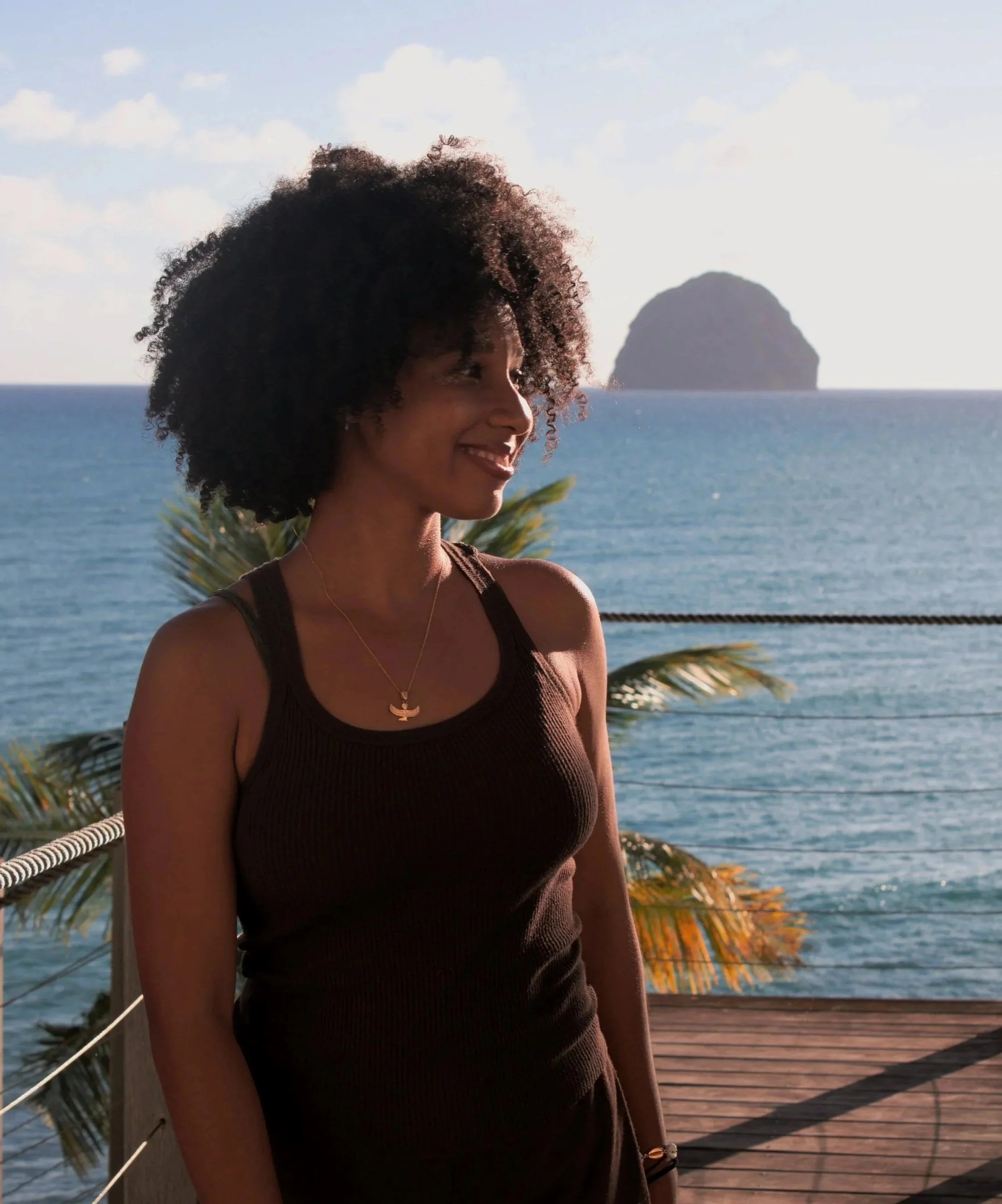 Femme souriante avec cheveux bouclés, portant un collier en or et une robe marron, se tenant près de la mer avec une île en arrière-plan.