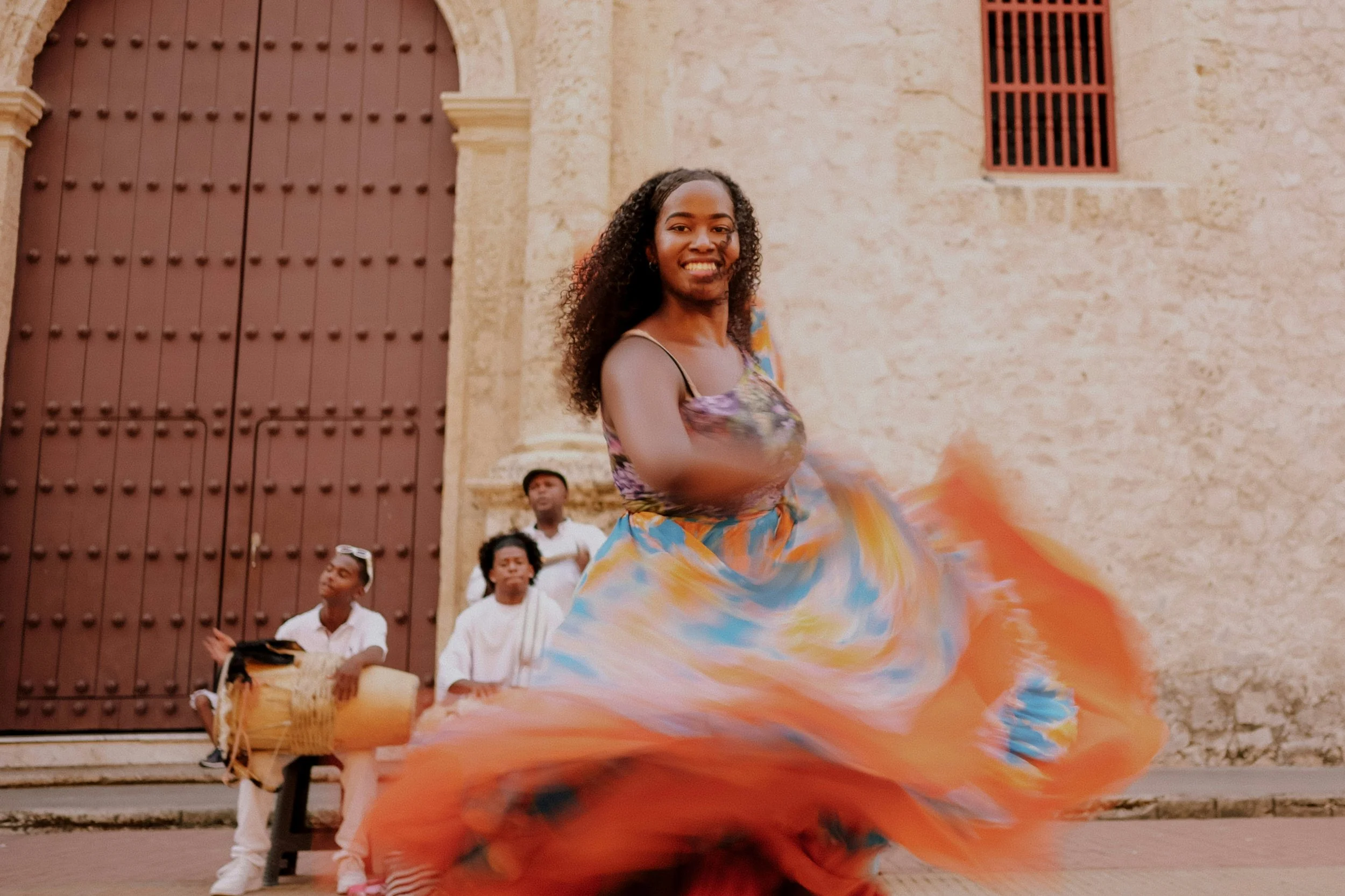 Une femme danse avec une robe fluide colorée devant un bâtiment ancien, entourée de musiciens.