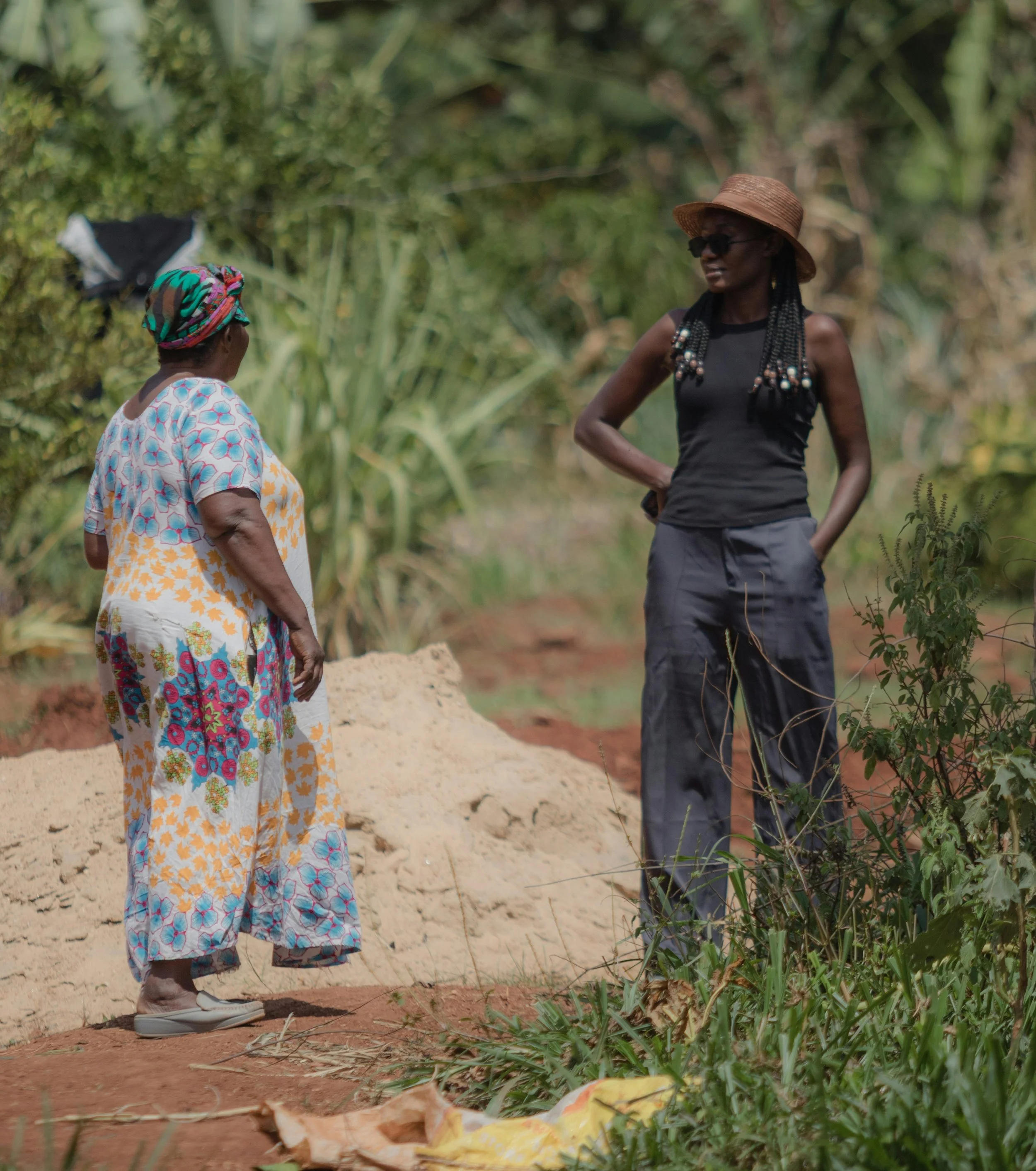 Deux femmes discutent dehors dans un environnement naturel, une d'elles porte une robe colorée et un foulard, l'autre porte un haut noir, une jupe longue grise, un chapeau en paille et des lunettes de soleil.